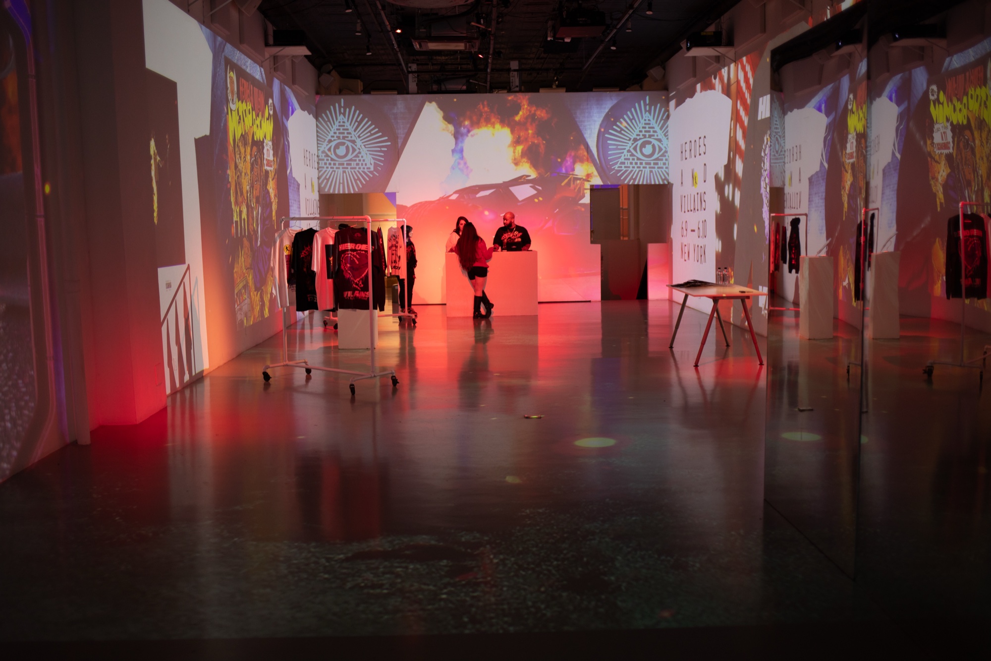A wide dark hall with clothing racks and a merch table under red projections of an eye and skyline