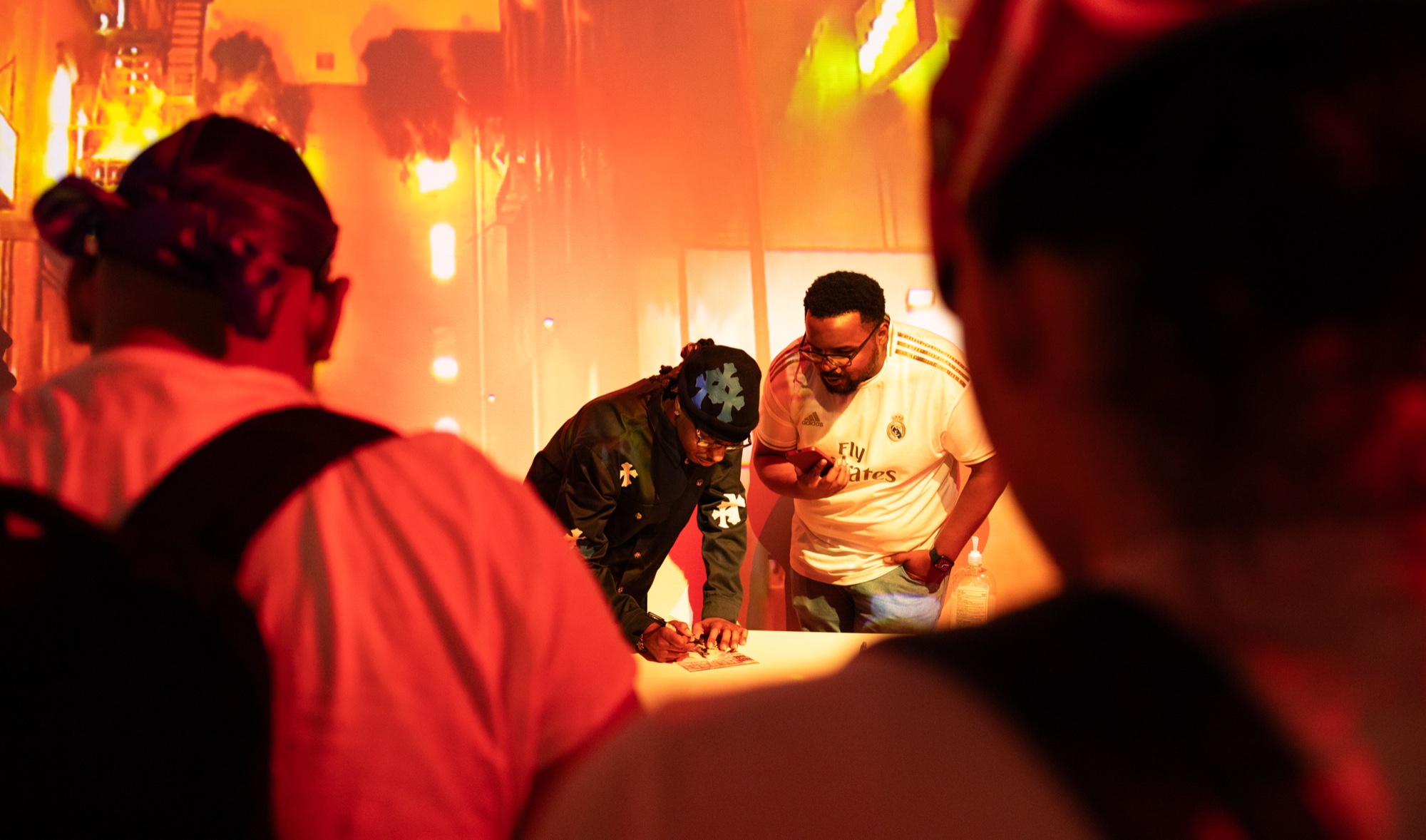 Two men in a cap and a white football jersey signing at a table under orange and red light