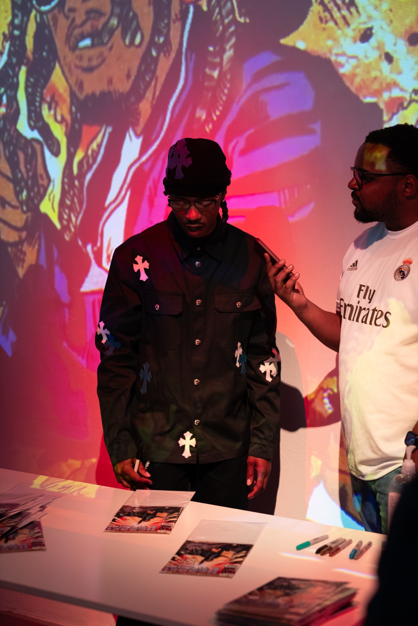 A man in a black shirt with cross patches looks down at signed prints, comic art projected behind him