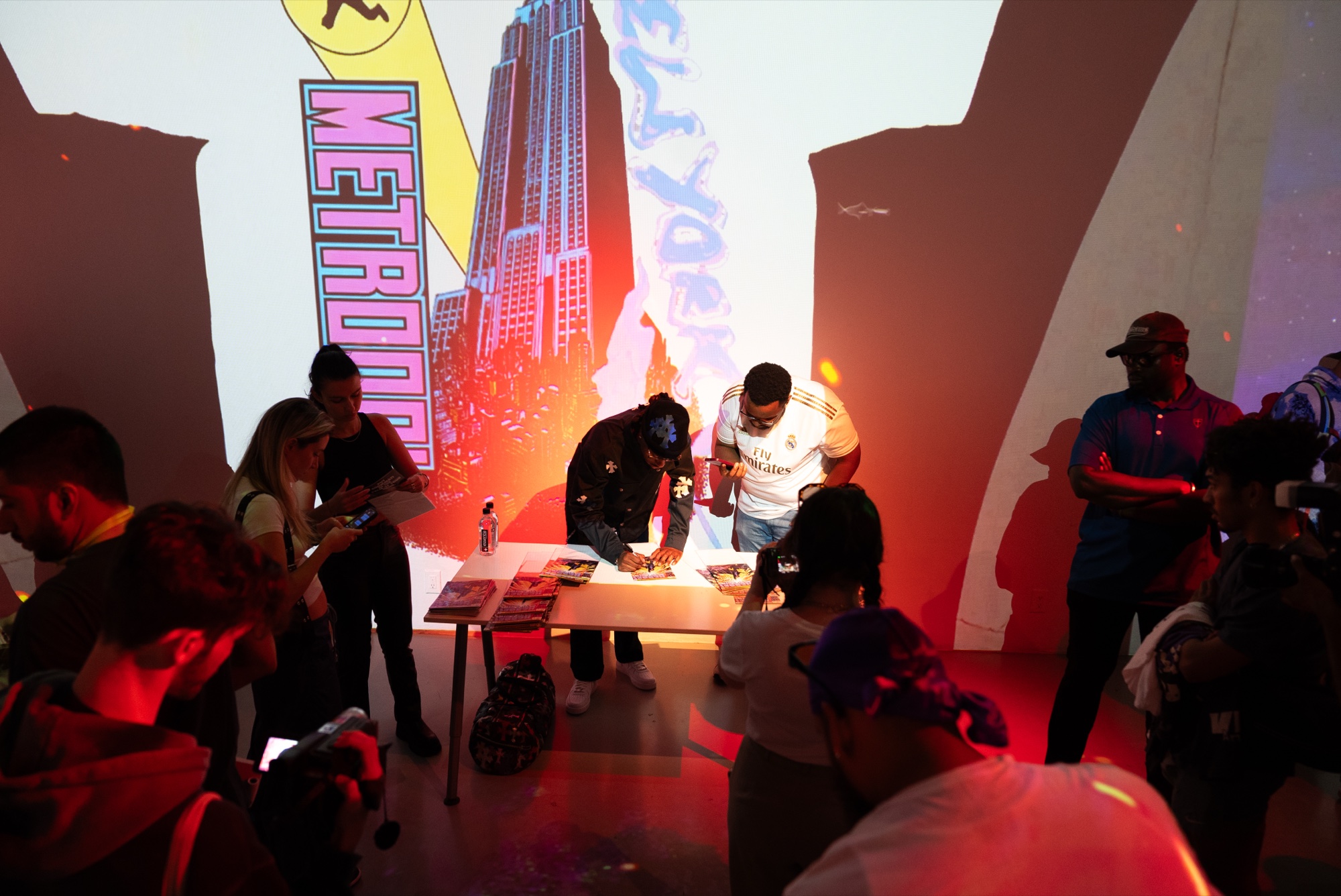 Guests crowd a signing table beneath a projected pink skyscraper and comic lettering in red light