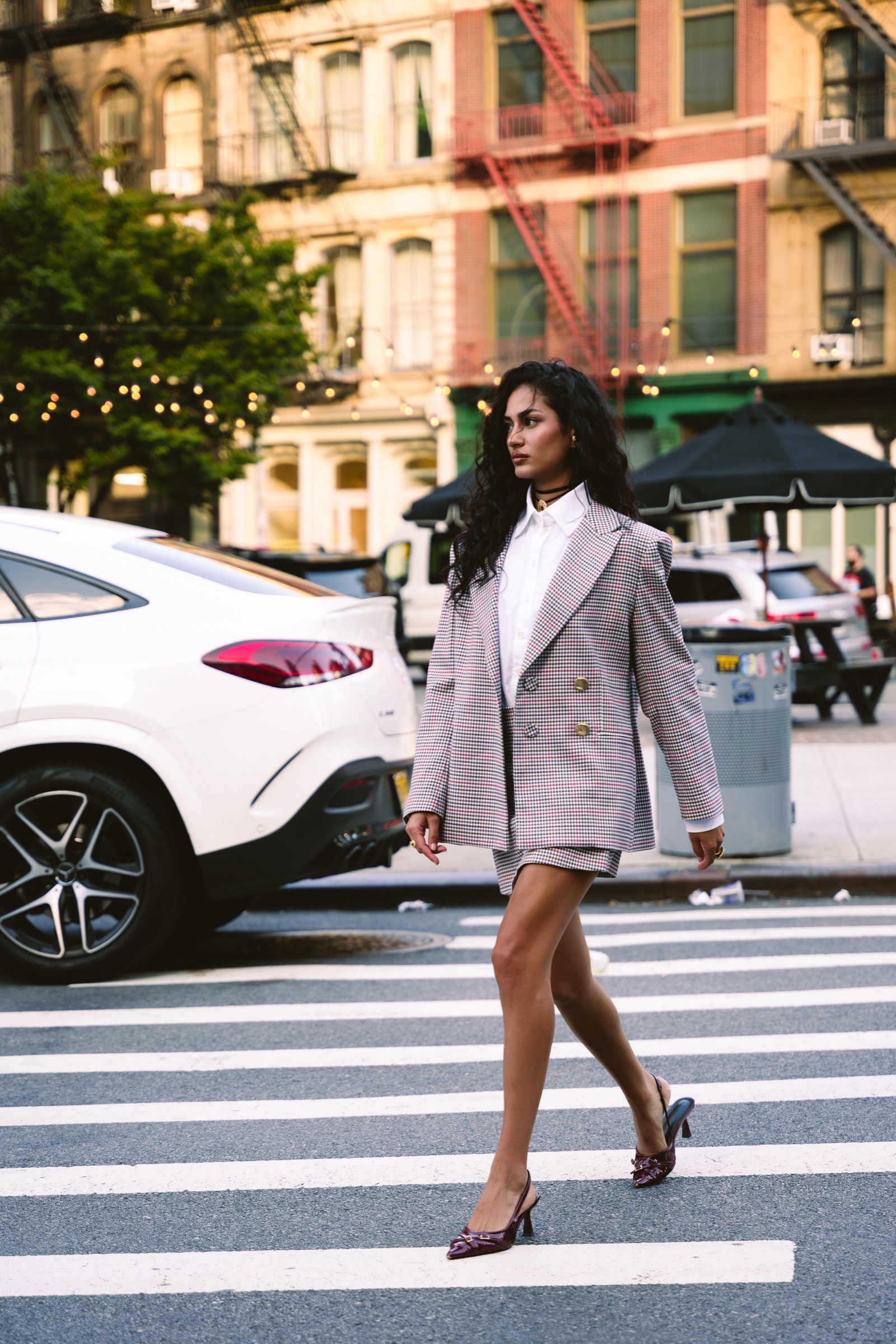 A model in a checked blazer and shorts strides across a crosswalk past a white car and red fire escapes
