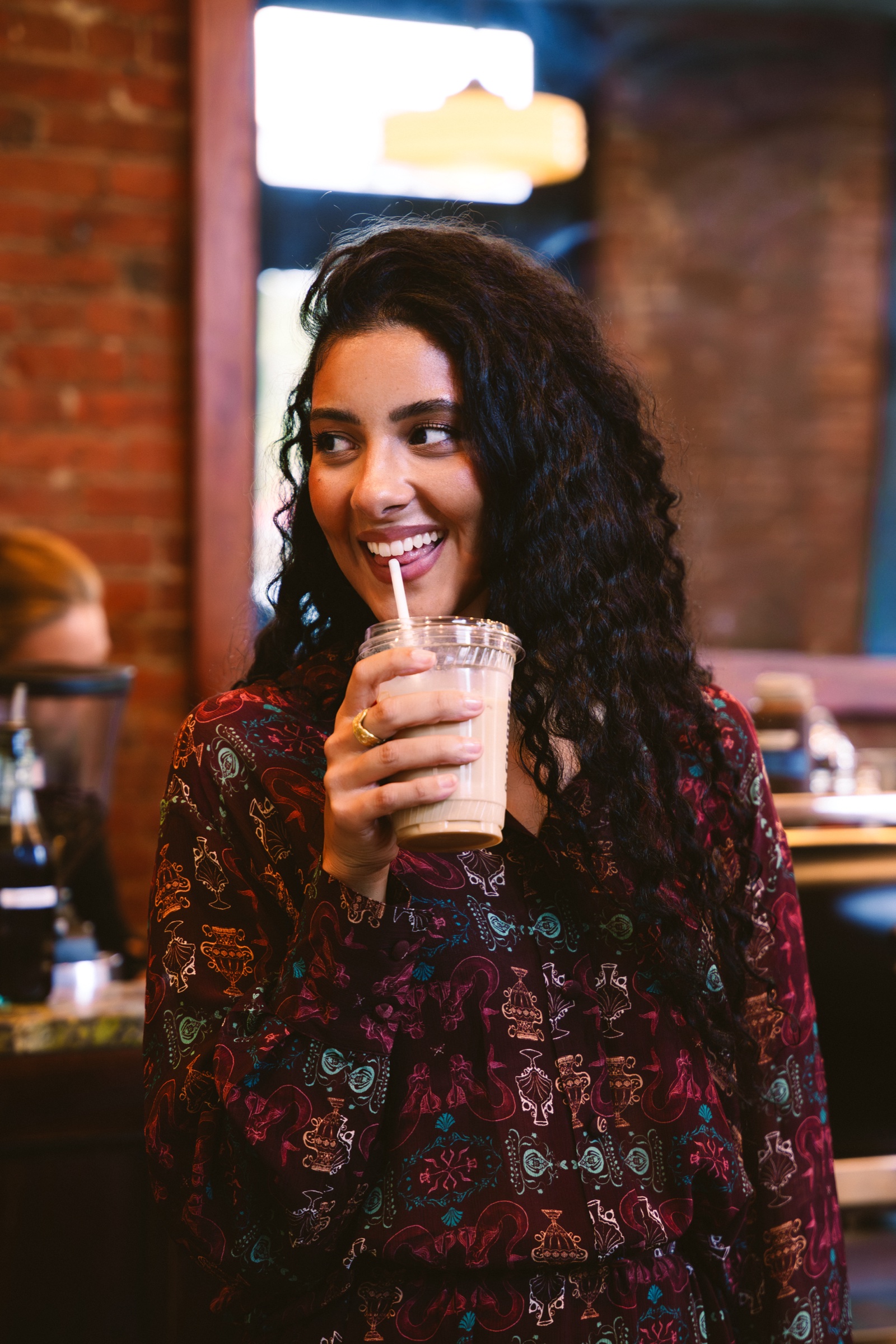 A smiling woman with long curly hair sips iced coffee through a straw in a brick walled cafe