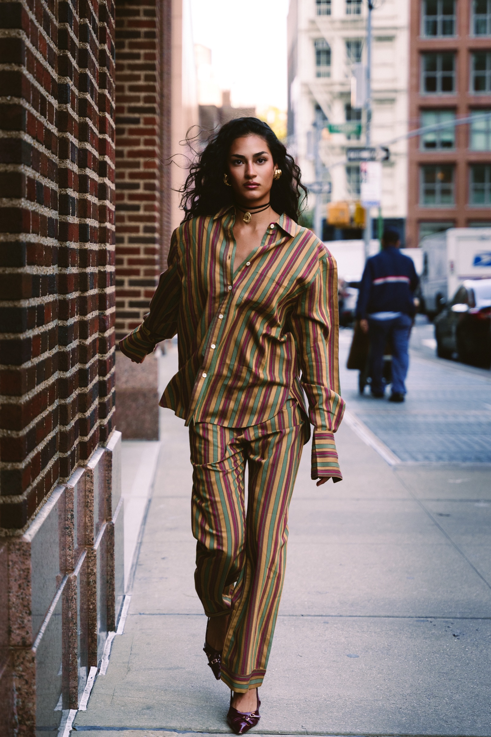 A model in a striped olive and burgundy shirt and trousers walks along a brick sidewalk in soft evening light