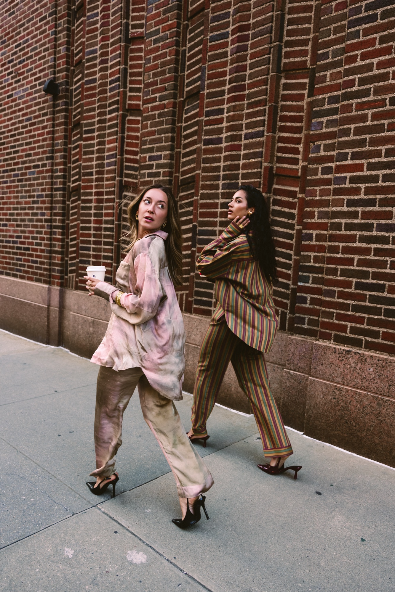 Two models in pink marbled and striped silk suits walk along a brick wall, one holding a coffee cup