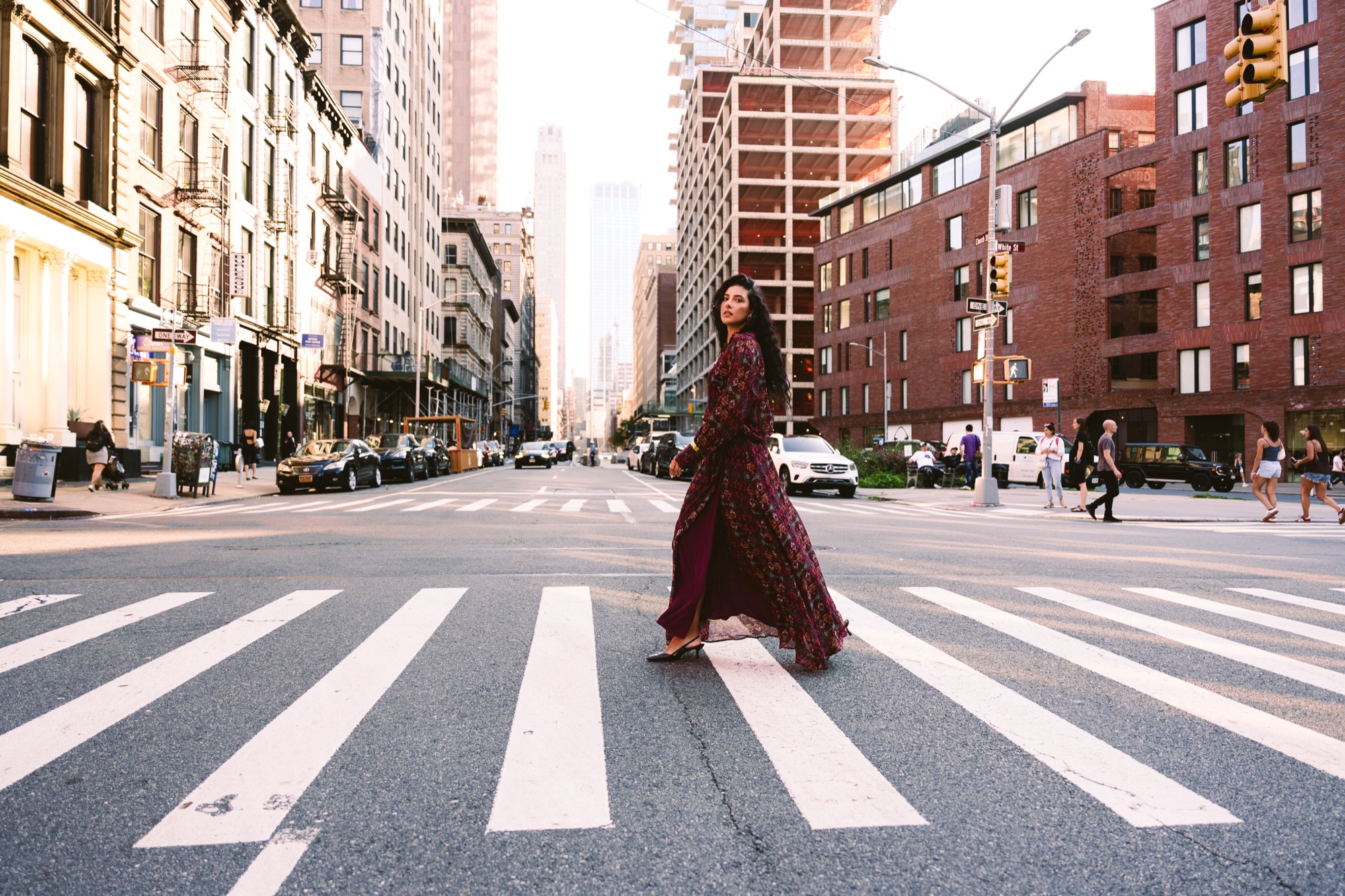 Woman in a long burgundy dress striding across a wide crosswalk between brick buildings in Manhattan