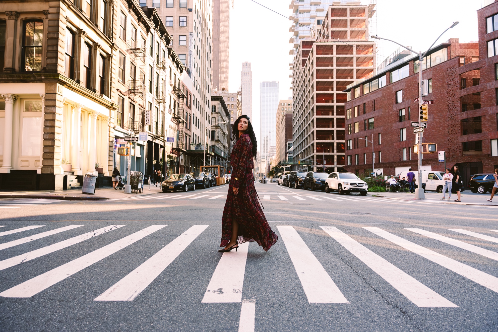 A model in a long dark red printed dress stands on a wide crosswalk between tall city buildings