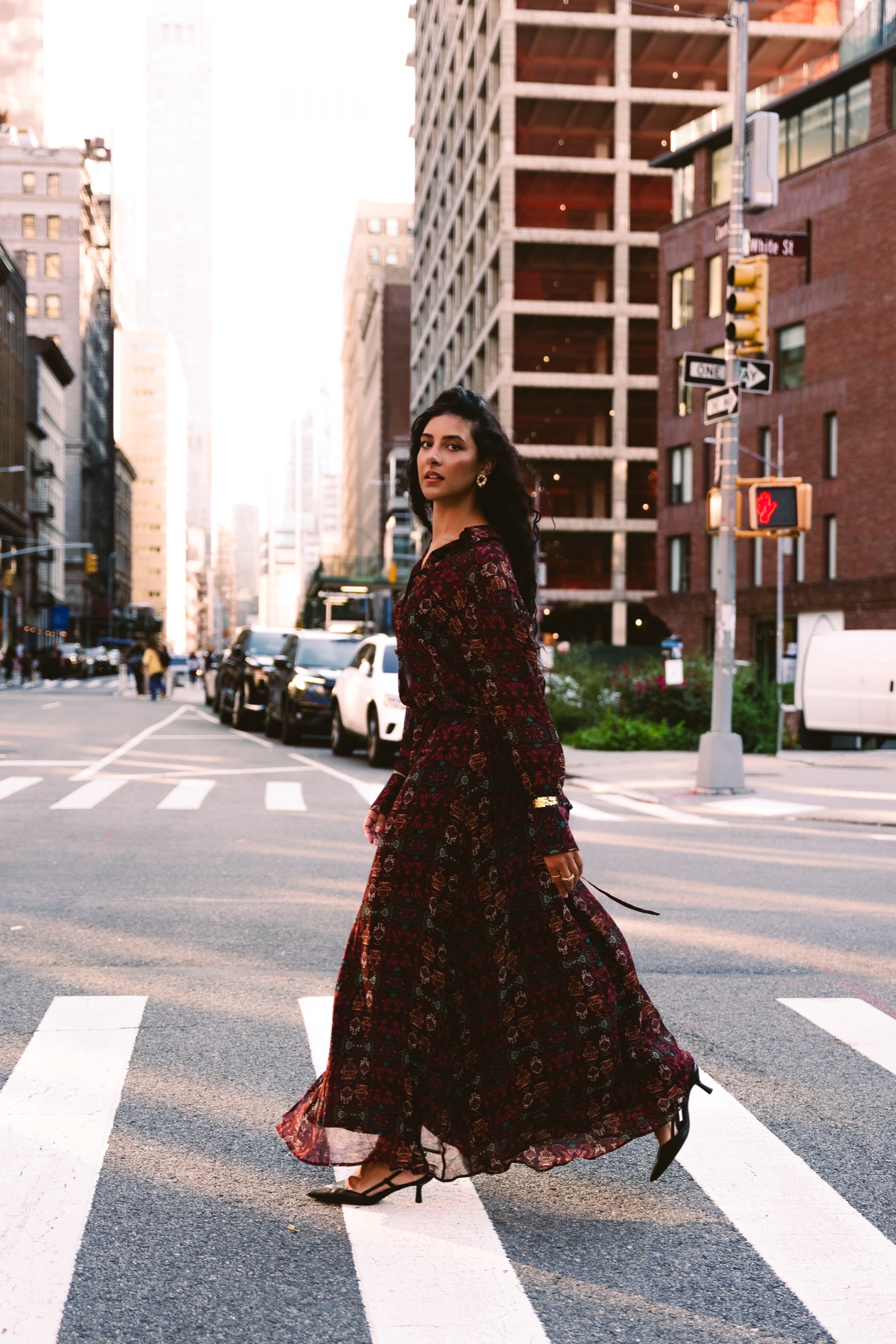 A model in a long dark floral dress crosses a Manhattan street on a crosswalk with tall buildings behind