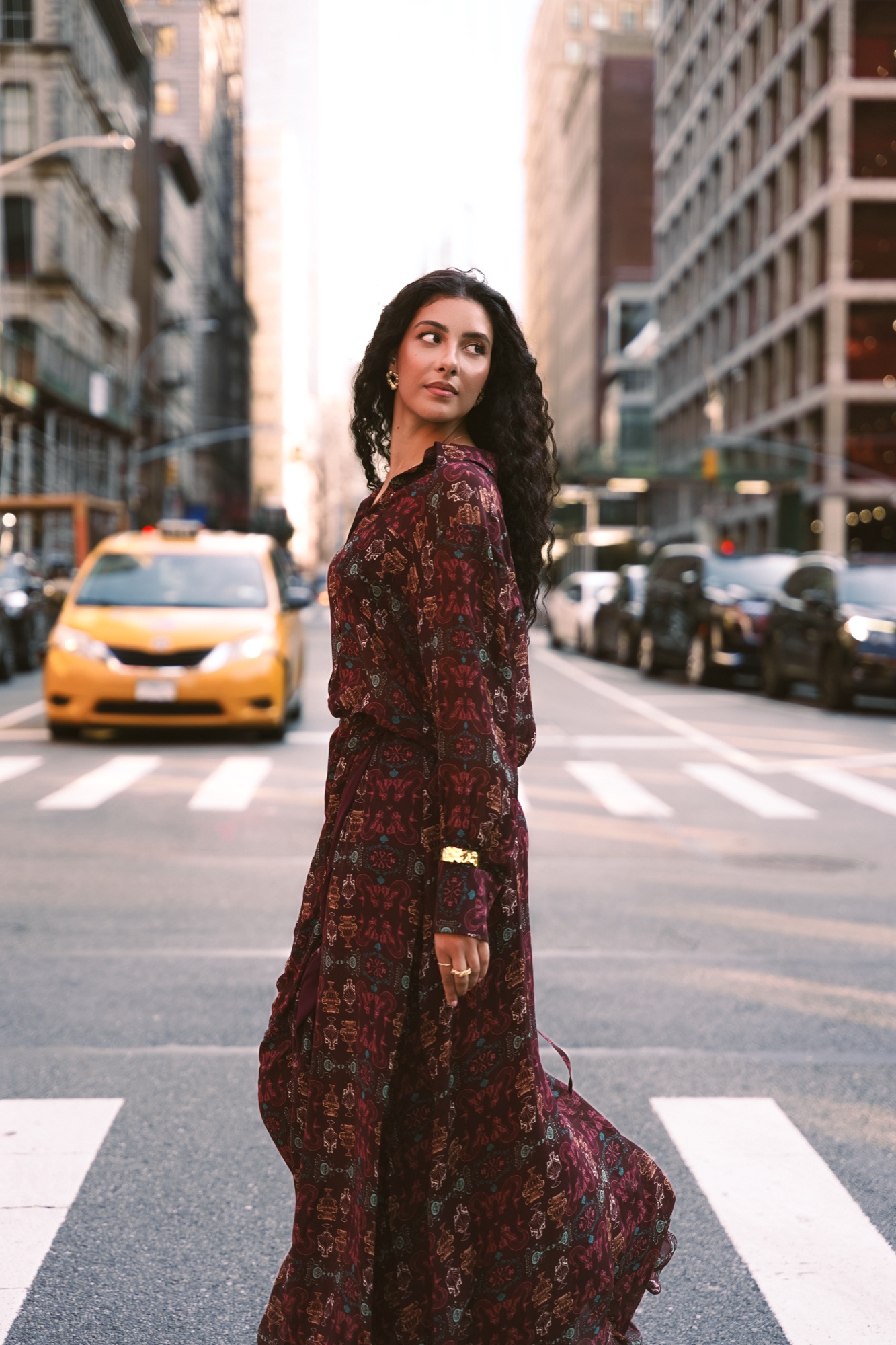 Woman in a patterned burgundy maxi dress glancing back on a crosswalk with a yellow taxi behind
