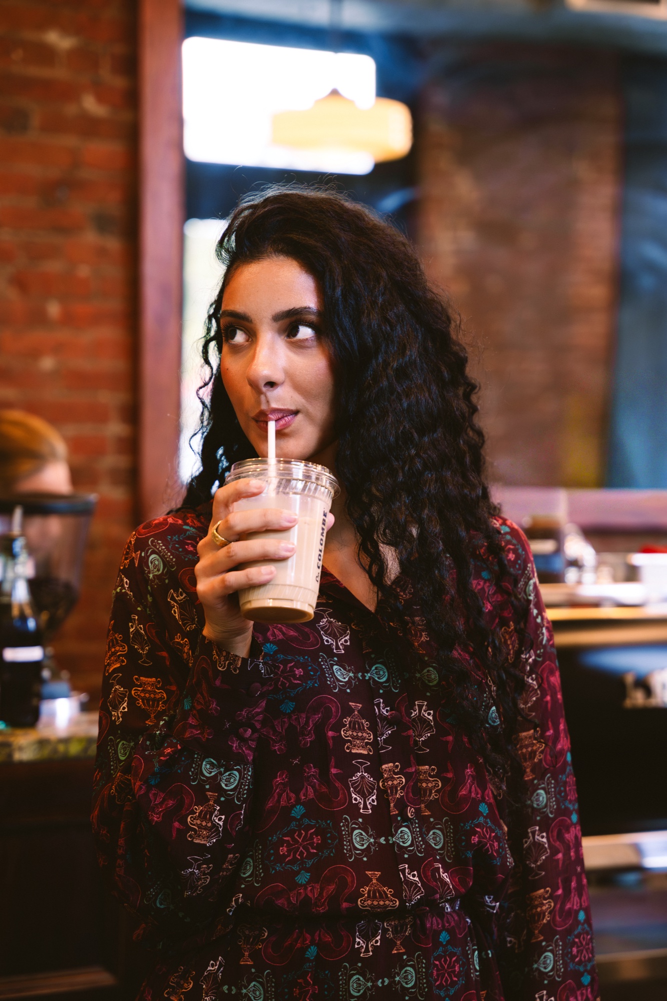 A woman with long curly hair in a burgundy print dress sips iced coffee in a brick walled cafe