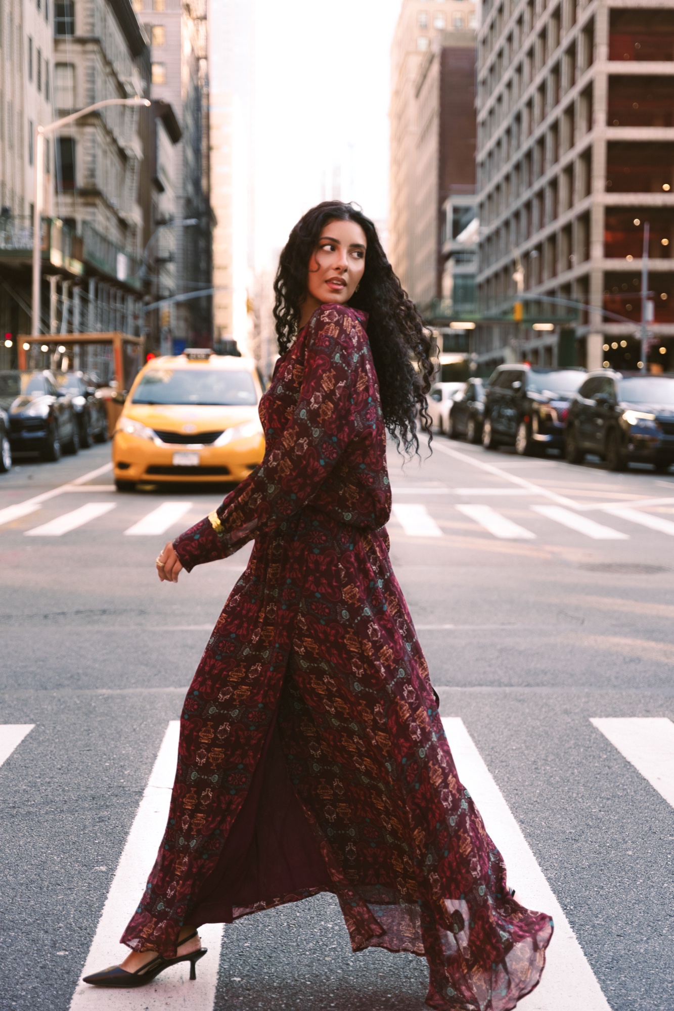 A woman in a flowing burgundy dress looks back over her shoulder on a crosswalk with a yellow taxi behind