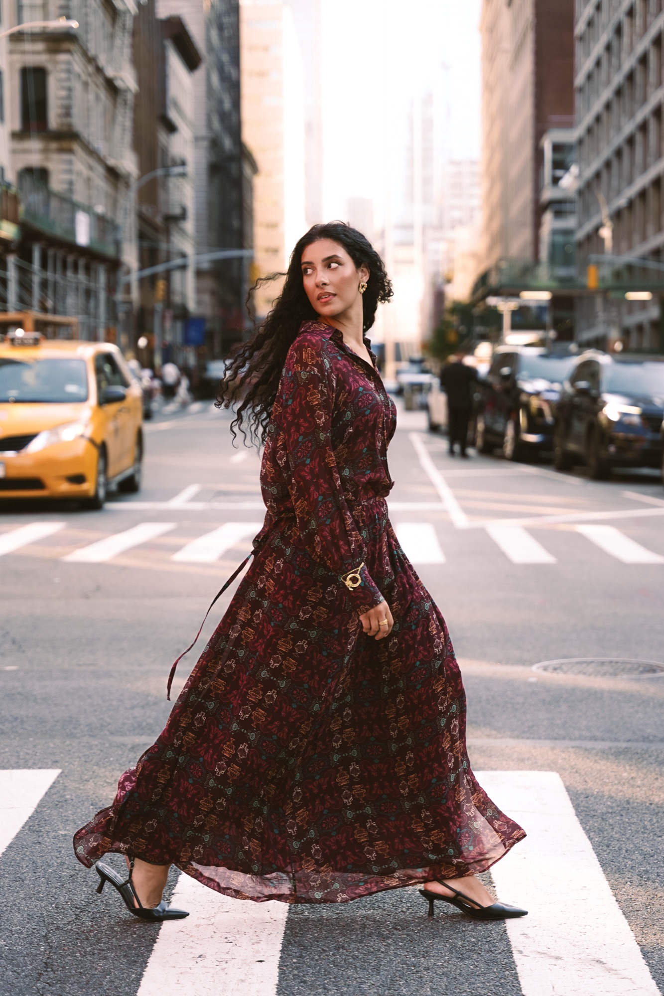 A model in a flowing dark floral dress strides across a crosswalk past a yellow taxi in low sunlight