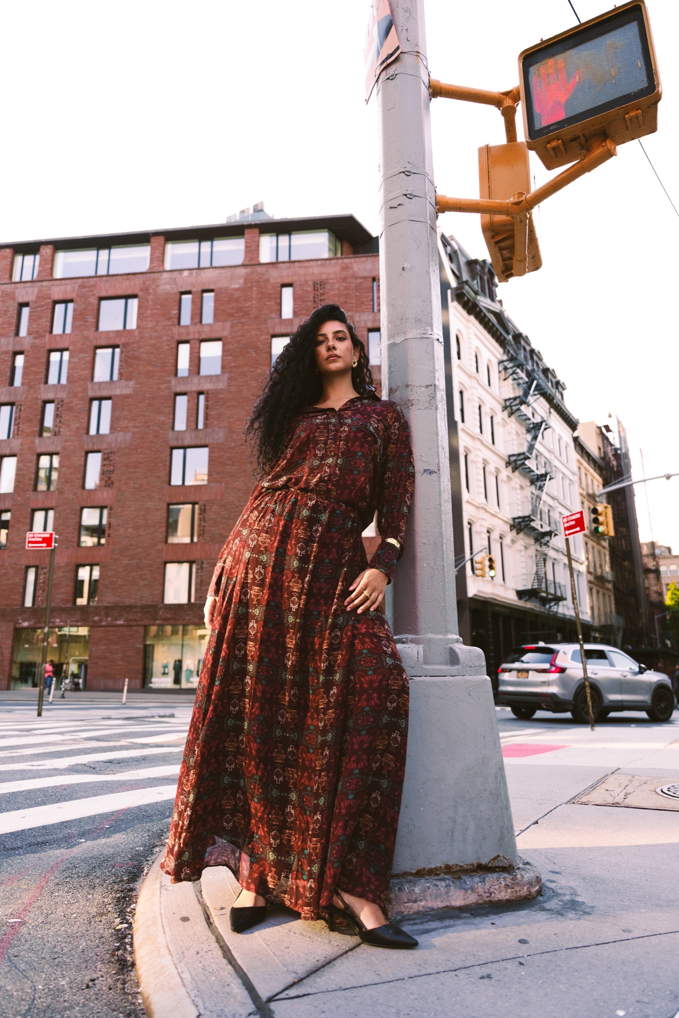 Woman in a long red patterned dress leaning against a traffic light pole beside a brick building