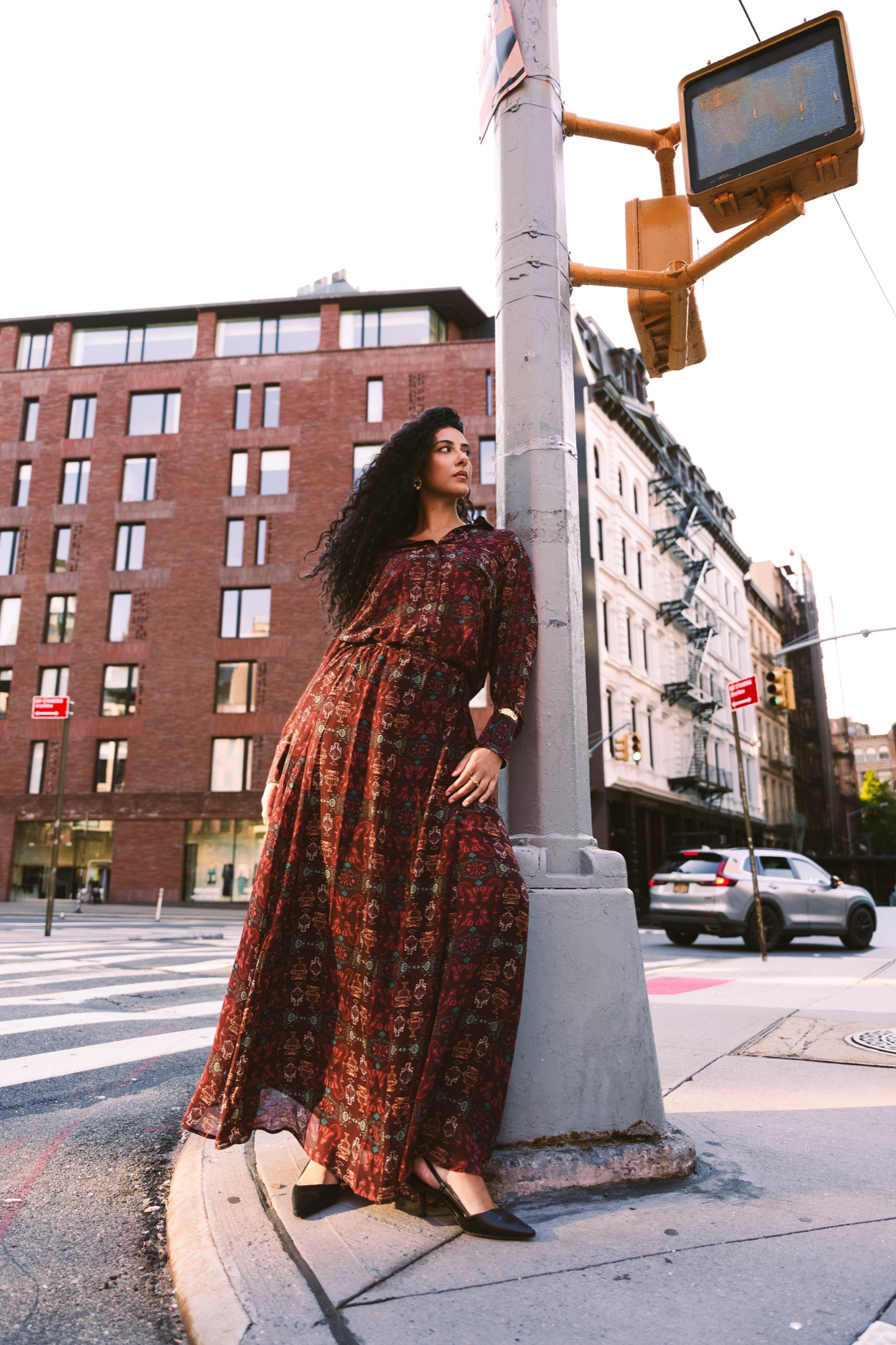 A model in a long red patterned dress leans against a traffic light pole beside a brick building