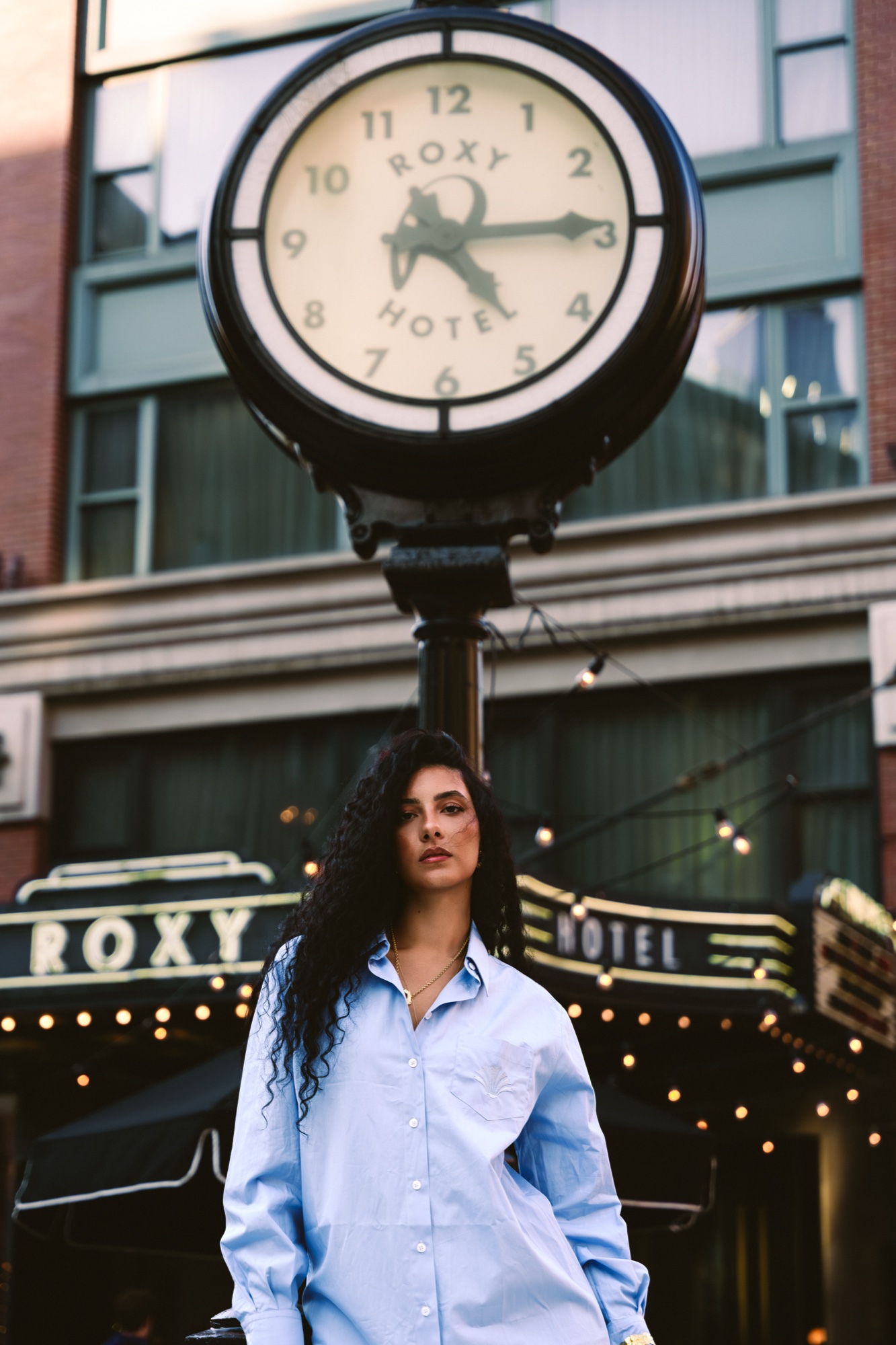 A woman in a pale blue shirt stands beneath the Roxy Hotel street clock and marquee lights