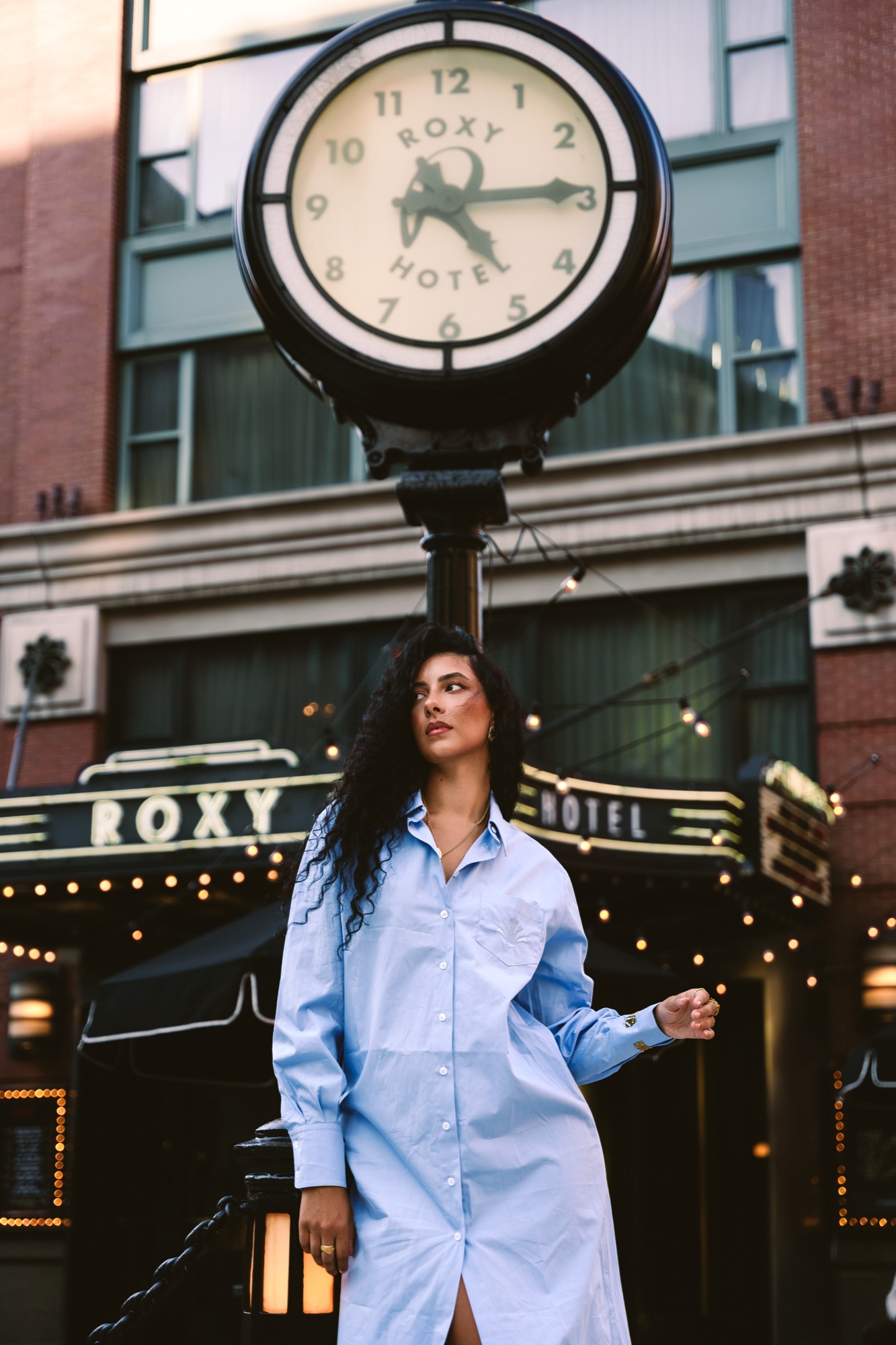 A model in an oversized pale blue shirt dress stands beneath the Roxy Hotel street clock and marquee
