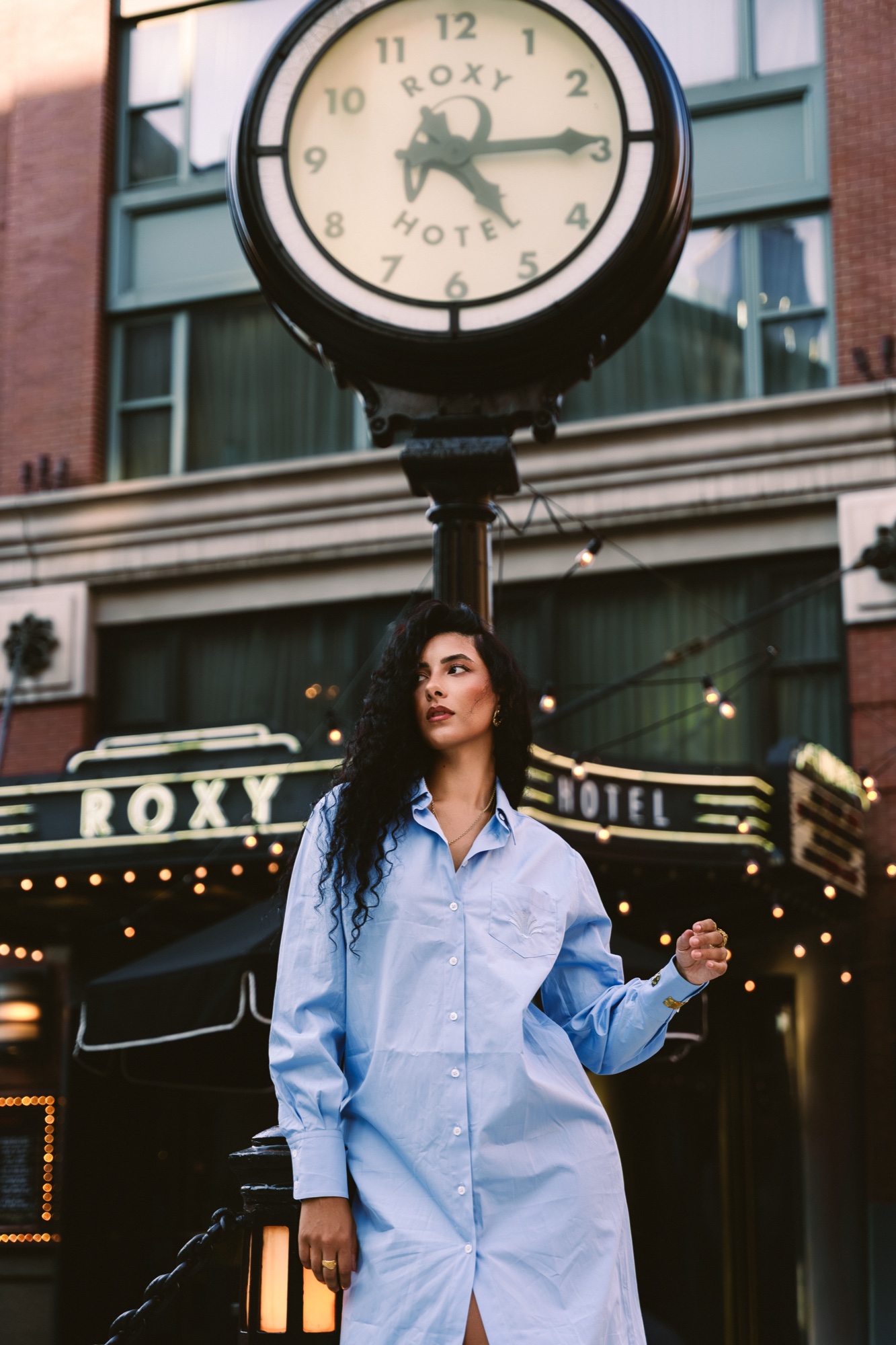 Woman in an oversized pale blue shirt dress beneath the Roxy Hotel clock and marquee lights