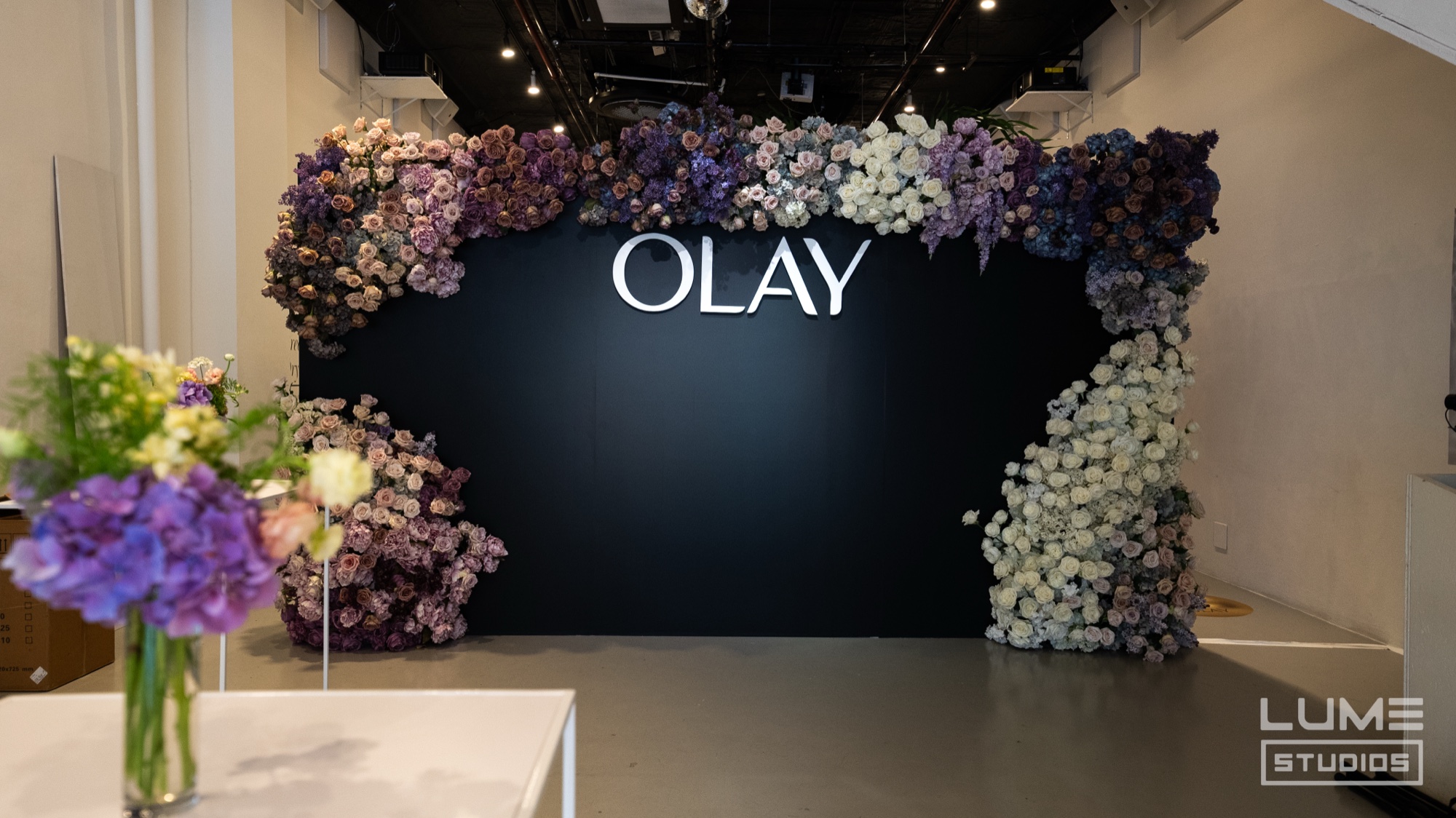 An arch of purple, lilac and white flowers frames a black wall printed with the Olay logo