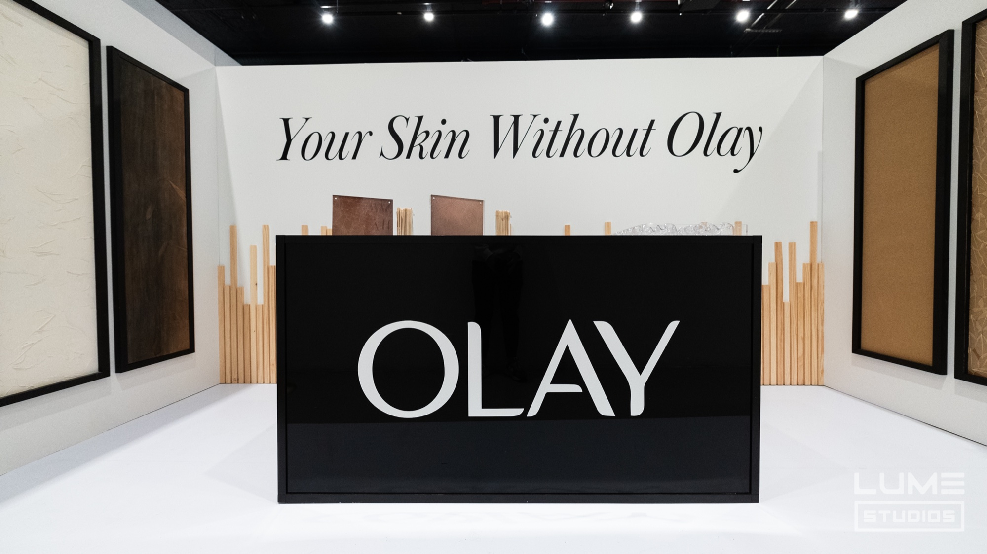 A black Olay sign box stands before a white wall reading Your Skin Without Olay, flanked by textured panels
