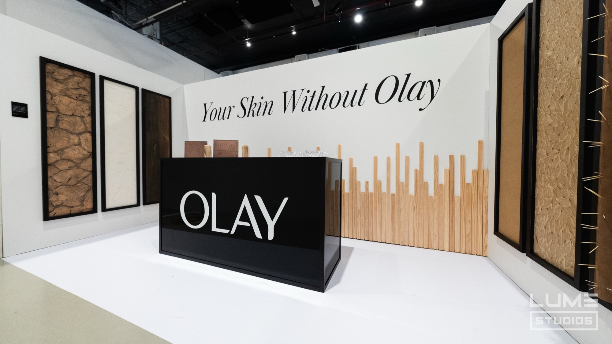 A black Olay counter stands before a white wall reading Your Skin Without Olay with panels of cracked textures