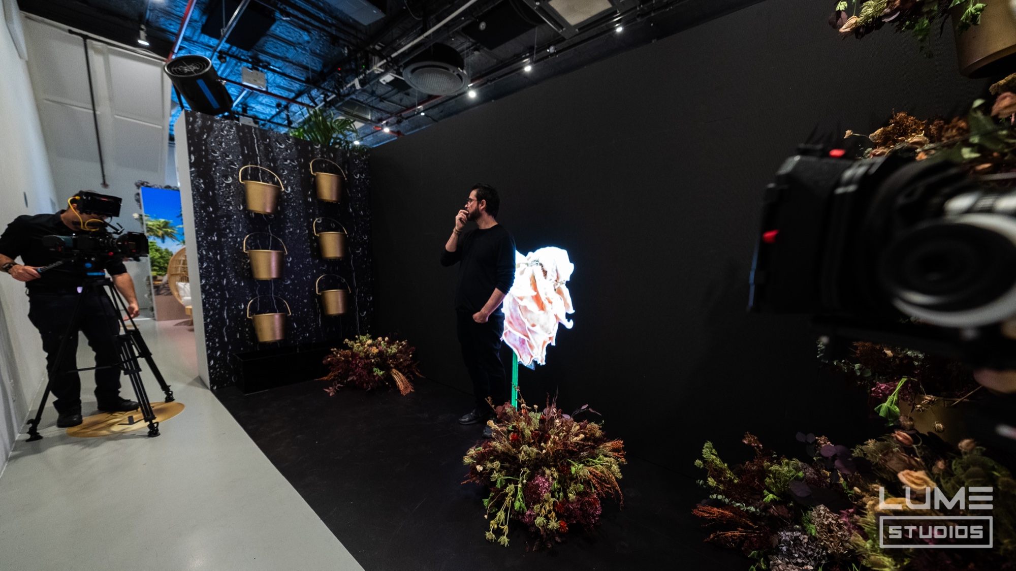 A camera operator films a person in black beside a glowing flower sculpture on a dark set with dried florals