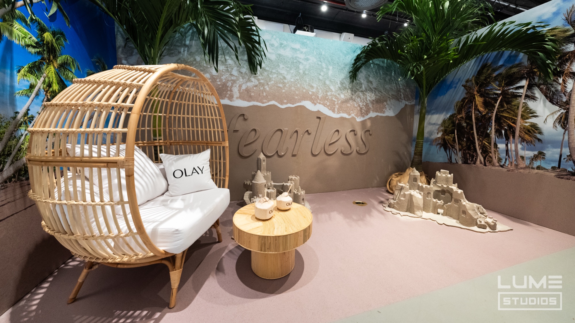 A rattan egg chair with an Olay cushion sits on pink sand beside a wall printed with the word Fearless