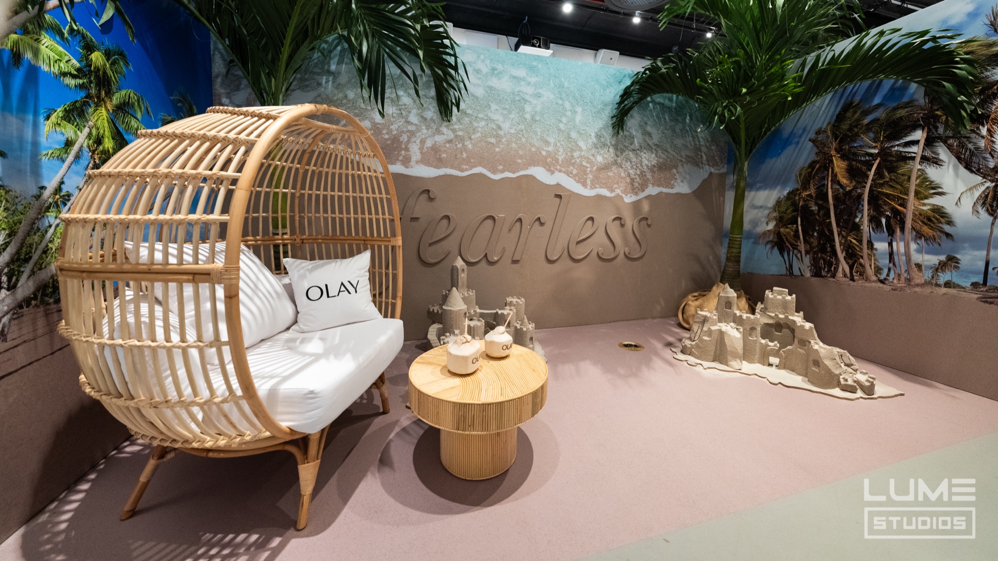 Rattan egg chair with an Olay pillow beside a sandcastle and palms before a beach mural reading fearless