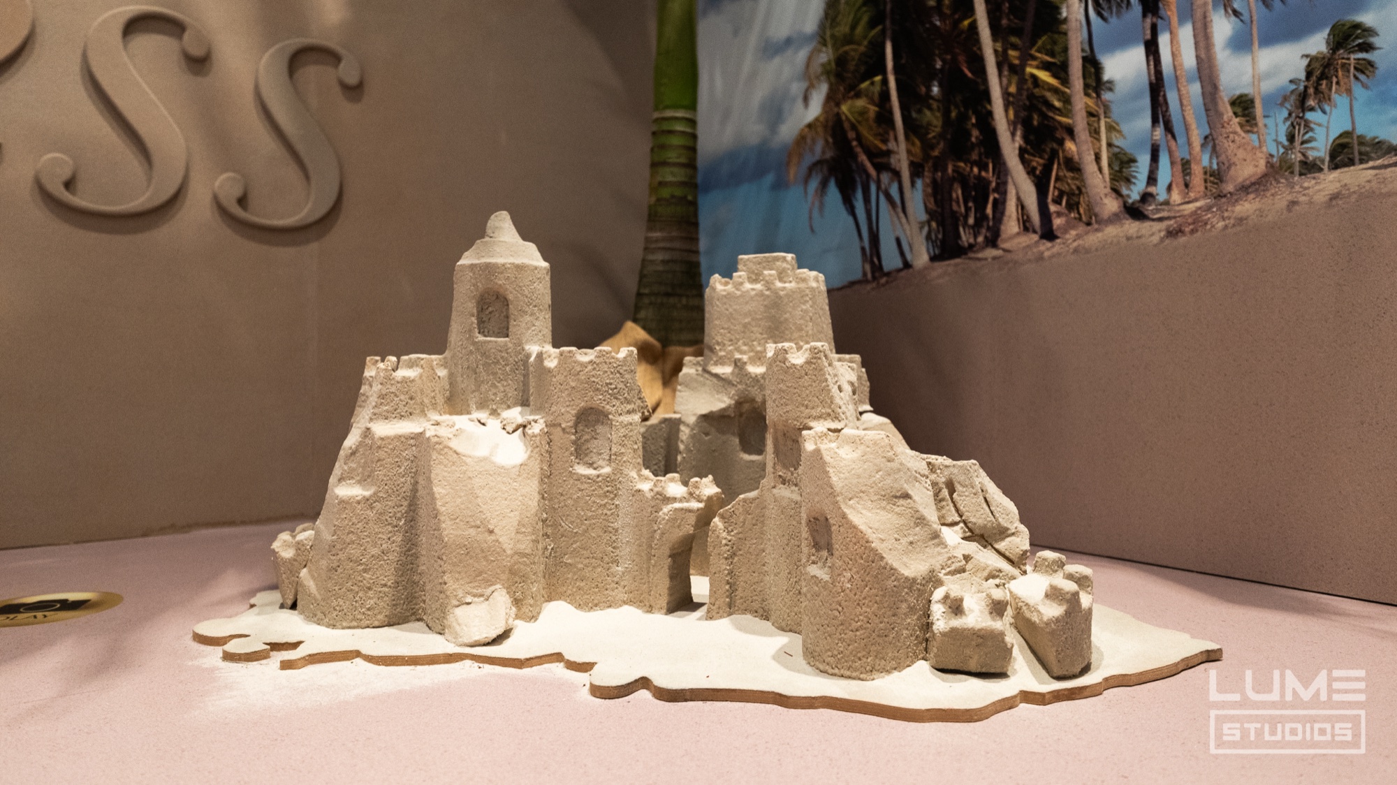 Detailed sand castle model on a pale pink surface in front of a tropical palm tree backdrop