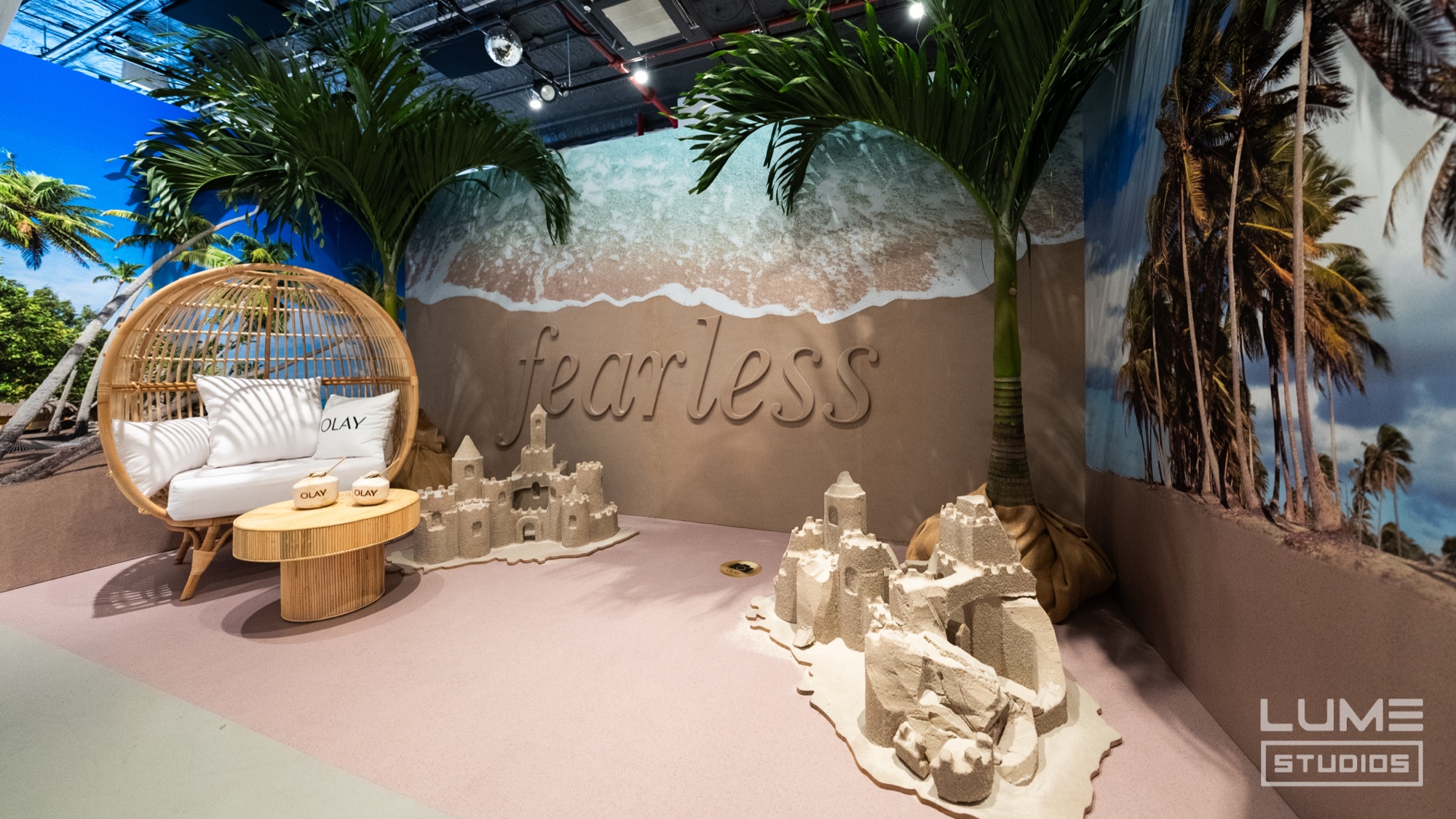 A beach set with sandcastles, palms and a rattan egg chair before a wall printed with the word Fearless