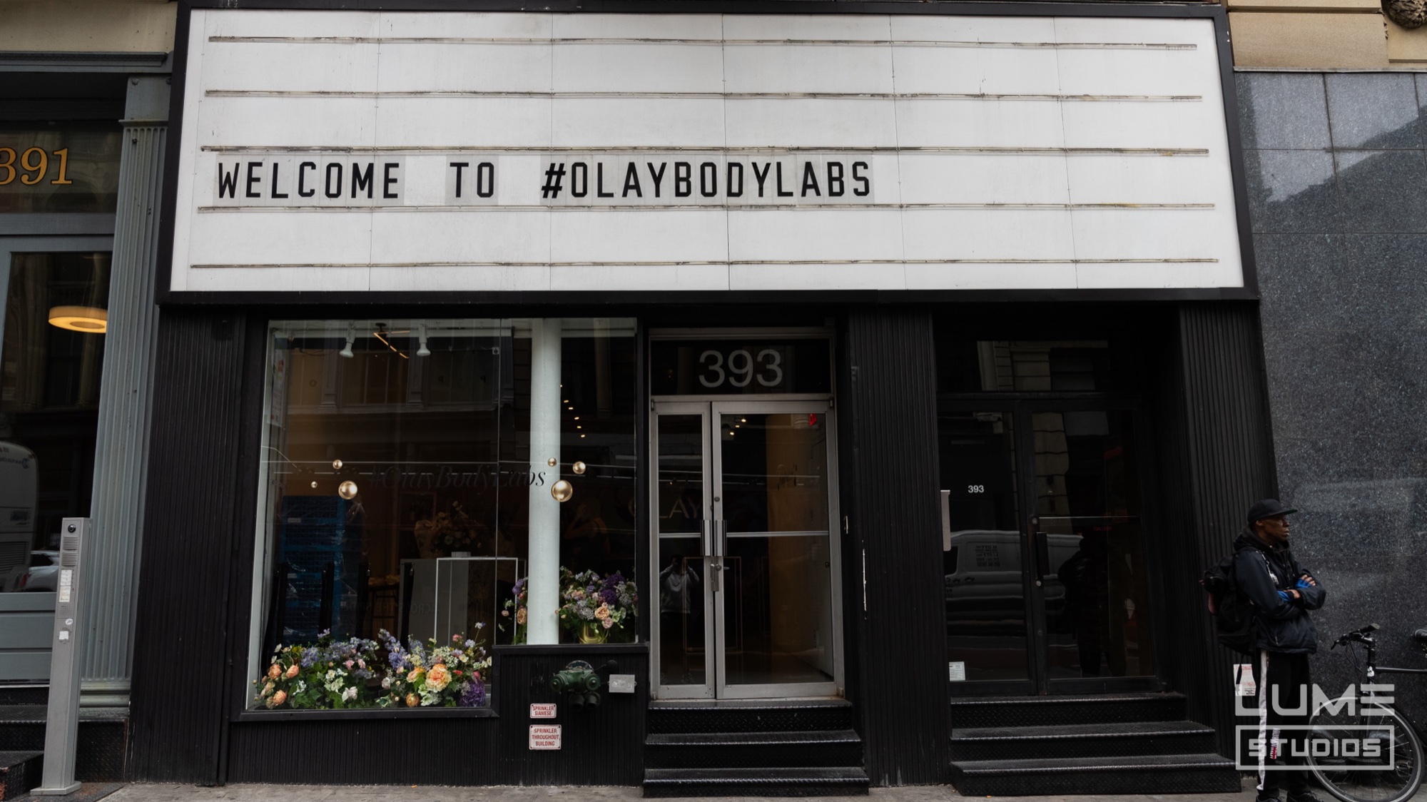 Black storefront marquee reading welcome to #olaybodylabs above a window filled with flowers