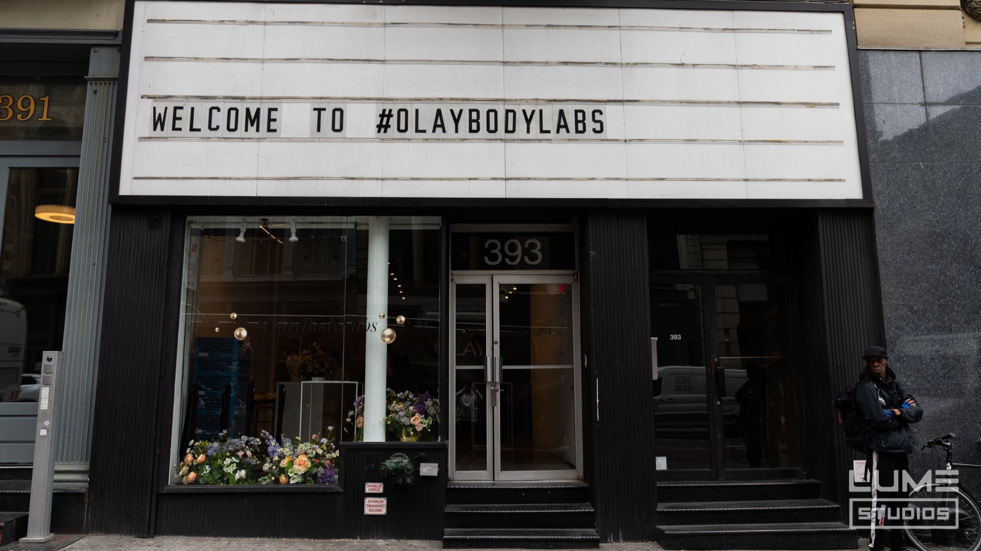 A marquee reads Welcome to #OlayBodyLabs above a black storefront with flowers in the window