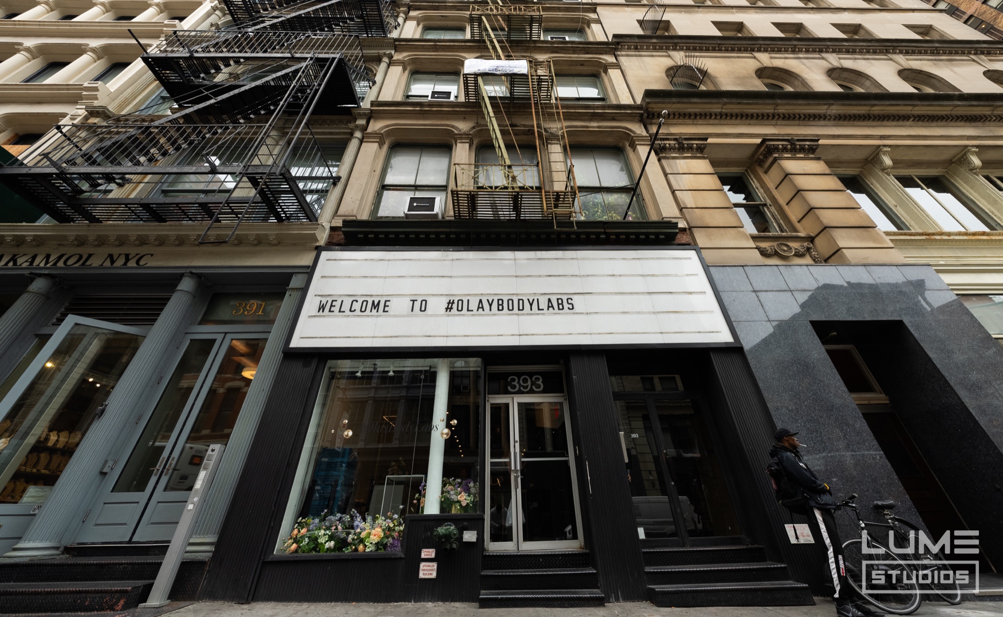 A storefront marquee reading Welcome to #OlayBodyLabs above a window of flowers on a cast iron block
