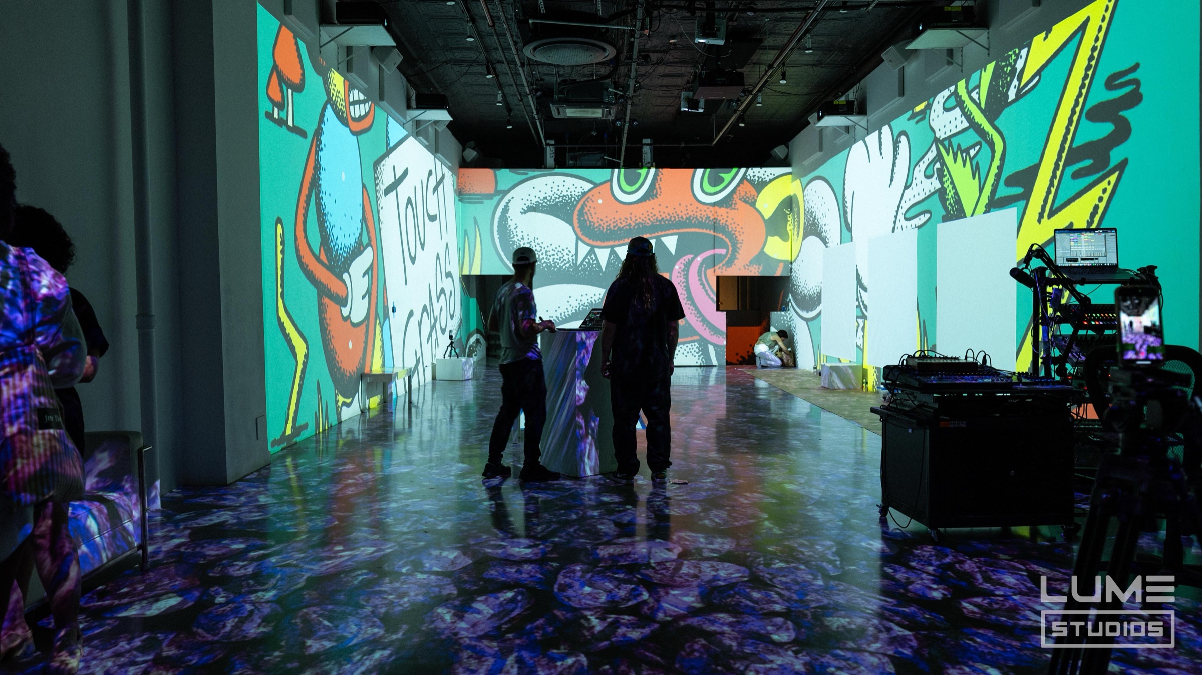 Guests stand among bold projected cartoon murals on a floor lit in purple at Paint with Pedro Baez