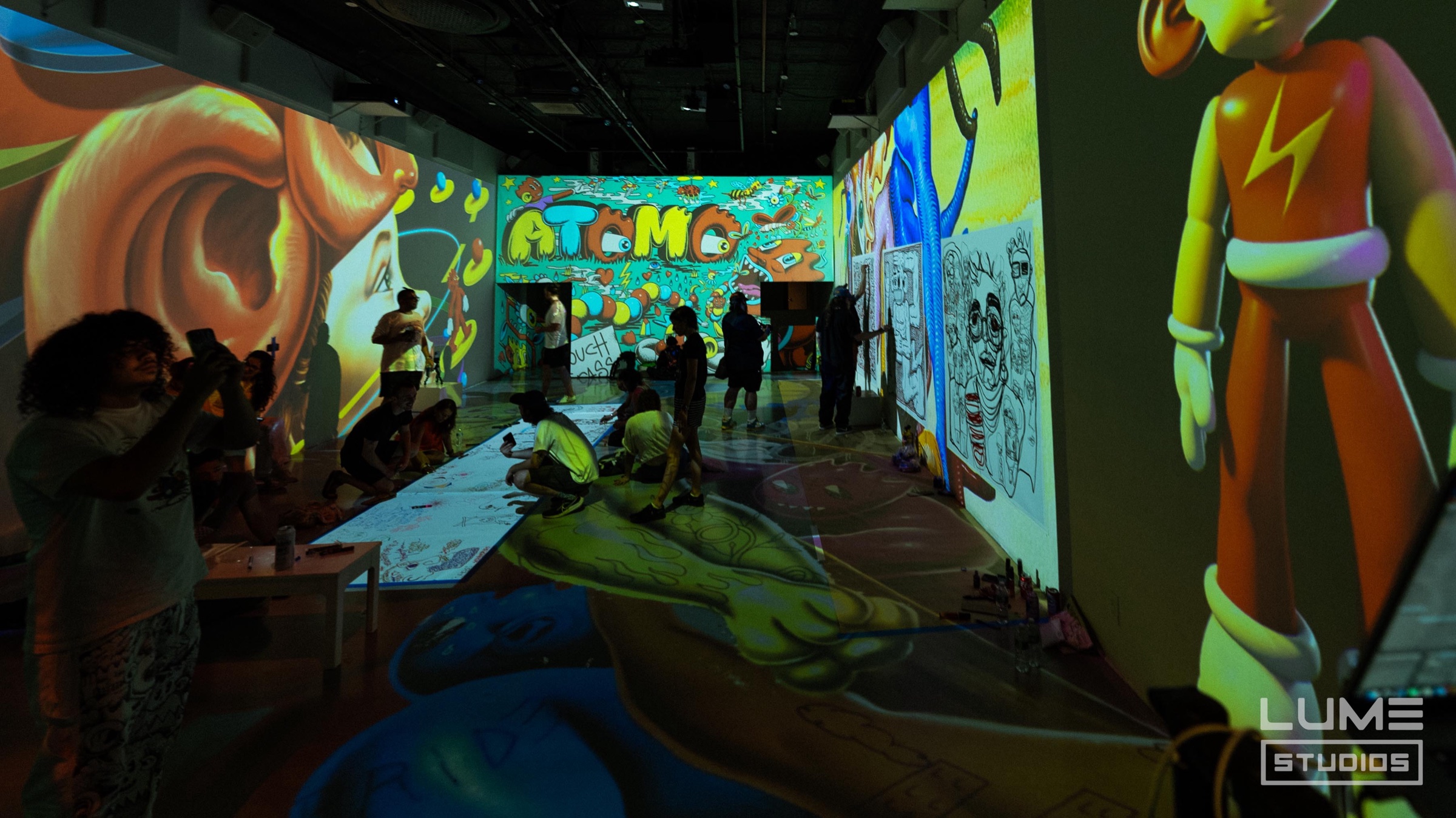 People drawing on floor canvases surrounded by vivid Atomo character projections during Paint with Pedro Baez