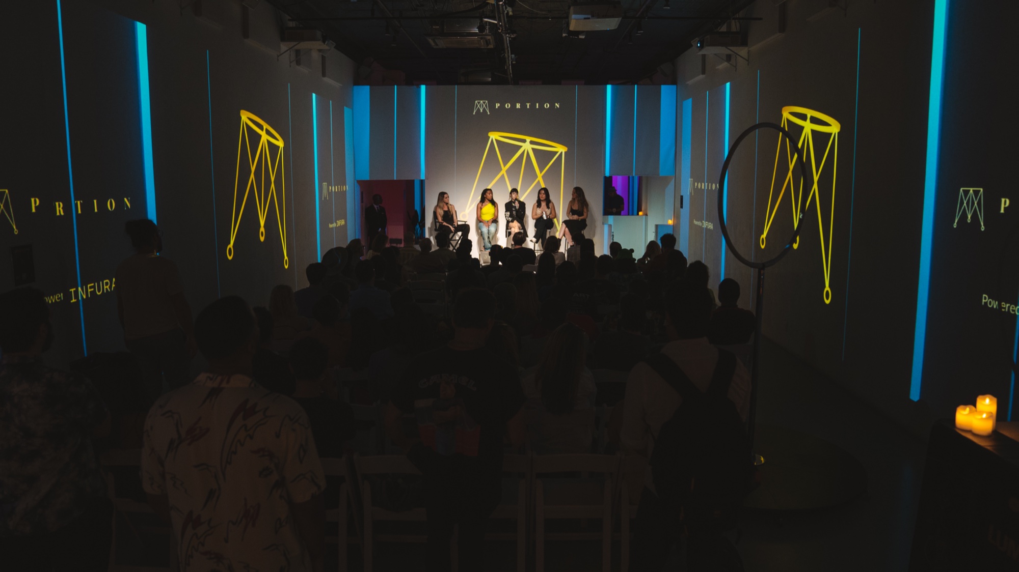A seated audience watches four performers on a stage flanked by yellow geometric shapes projected on the walls