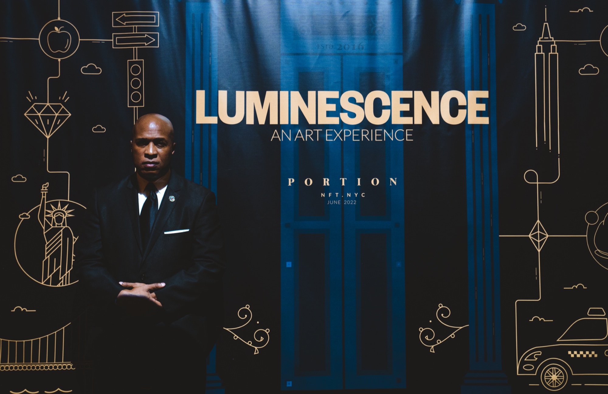 A man in a dark suit stands with arms folded beneath a Luminescence An Art Experience sign