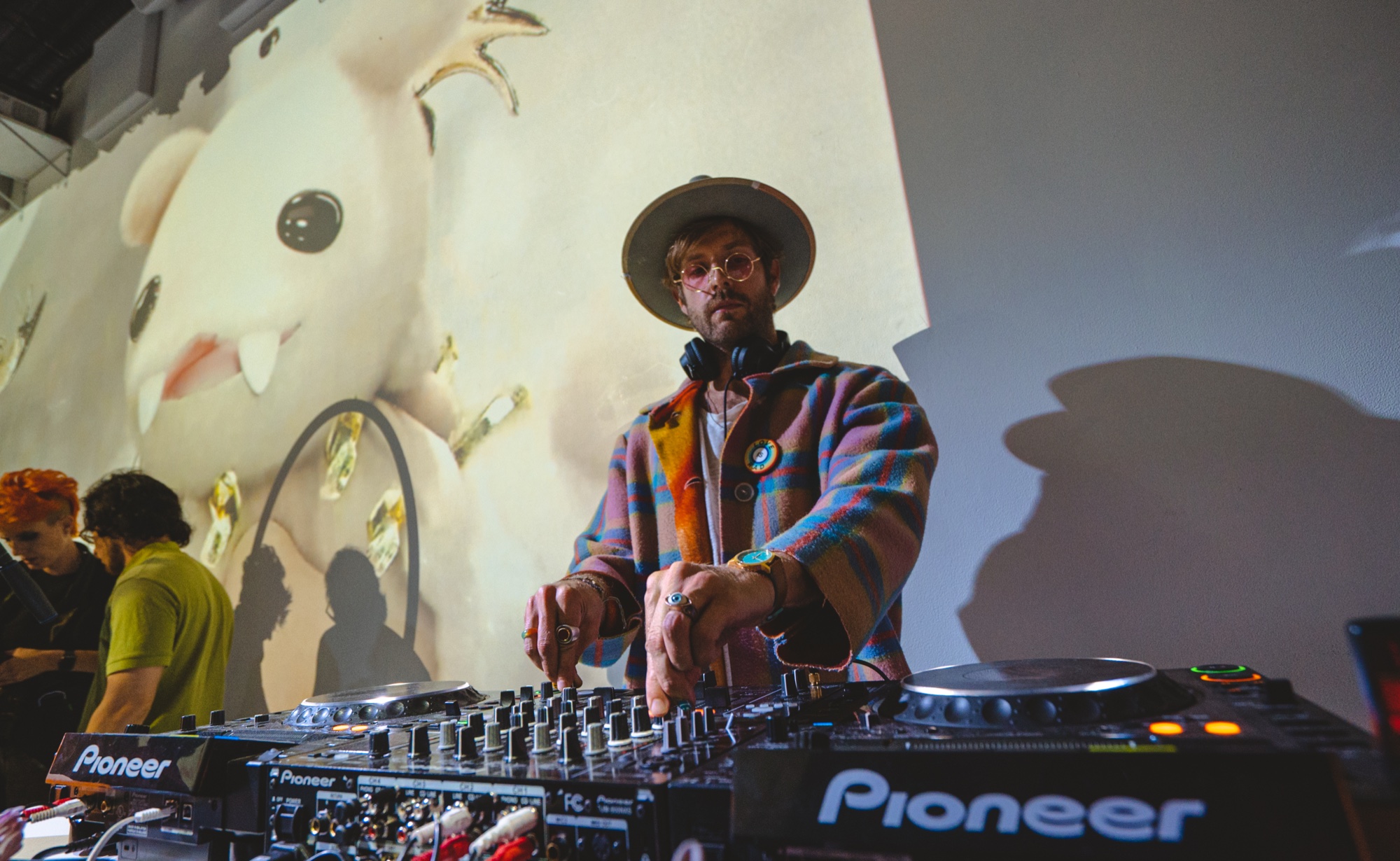 DJ in a wide brimmed hat at Pioneer decks before a projected cartoon creature on the wall