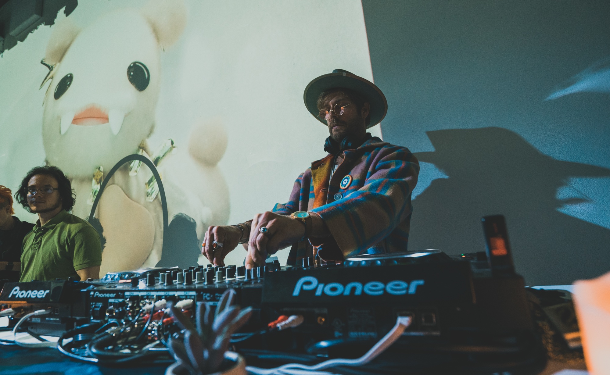 A DJ in a hat and plaid jacket works a Pioneer mixer before a projected cartoon creature in blue light