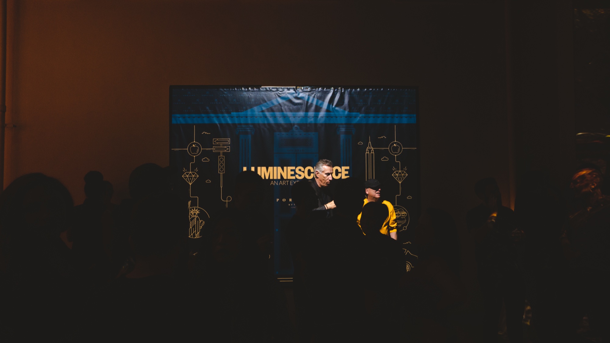 Dark room where silhouetted guests face a blue screen with Luminescence lettering and two figures at a table