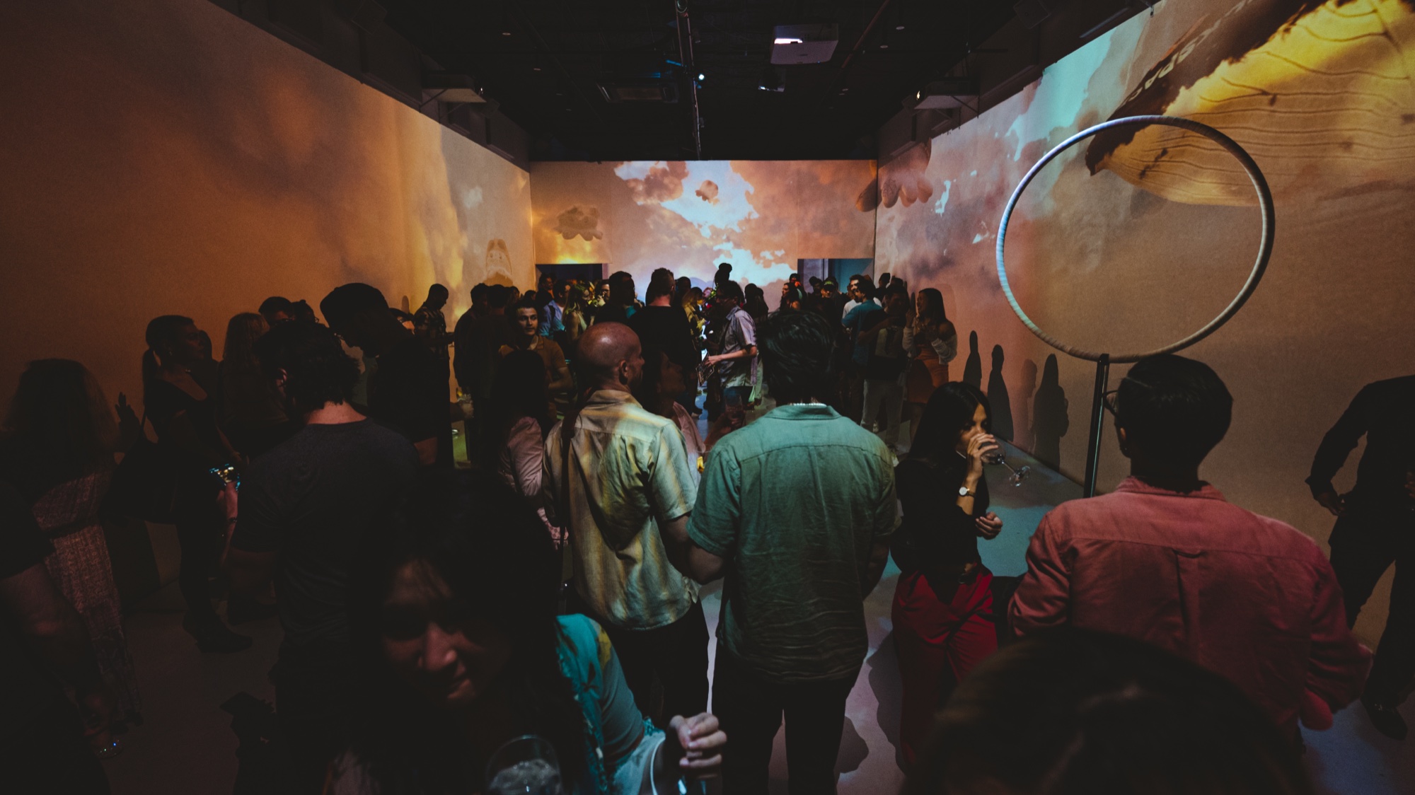 A crowd fills a dark room with orange and gold projections on the walls and a round mirror at right
