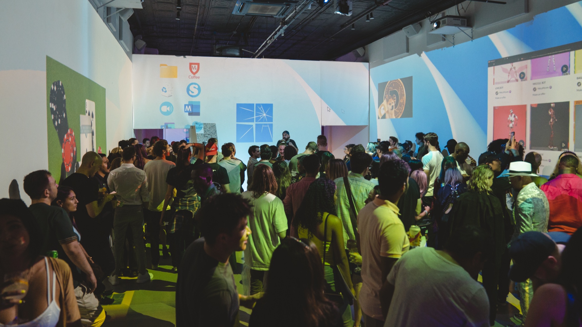 Crowd facing a speaker in a white gallery lit with green and blue projections of app icons