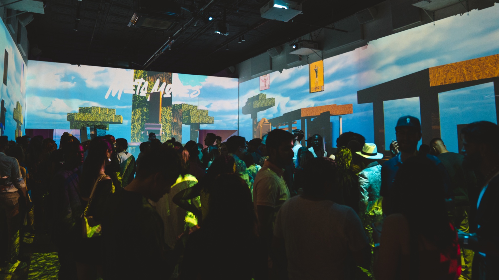 A crowd fills a room where a pixelated video game landscape with clouds is projected on the walls