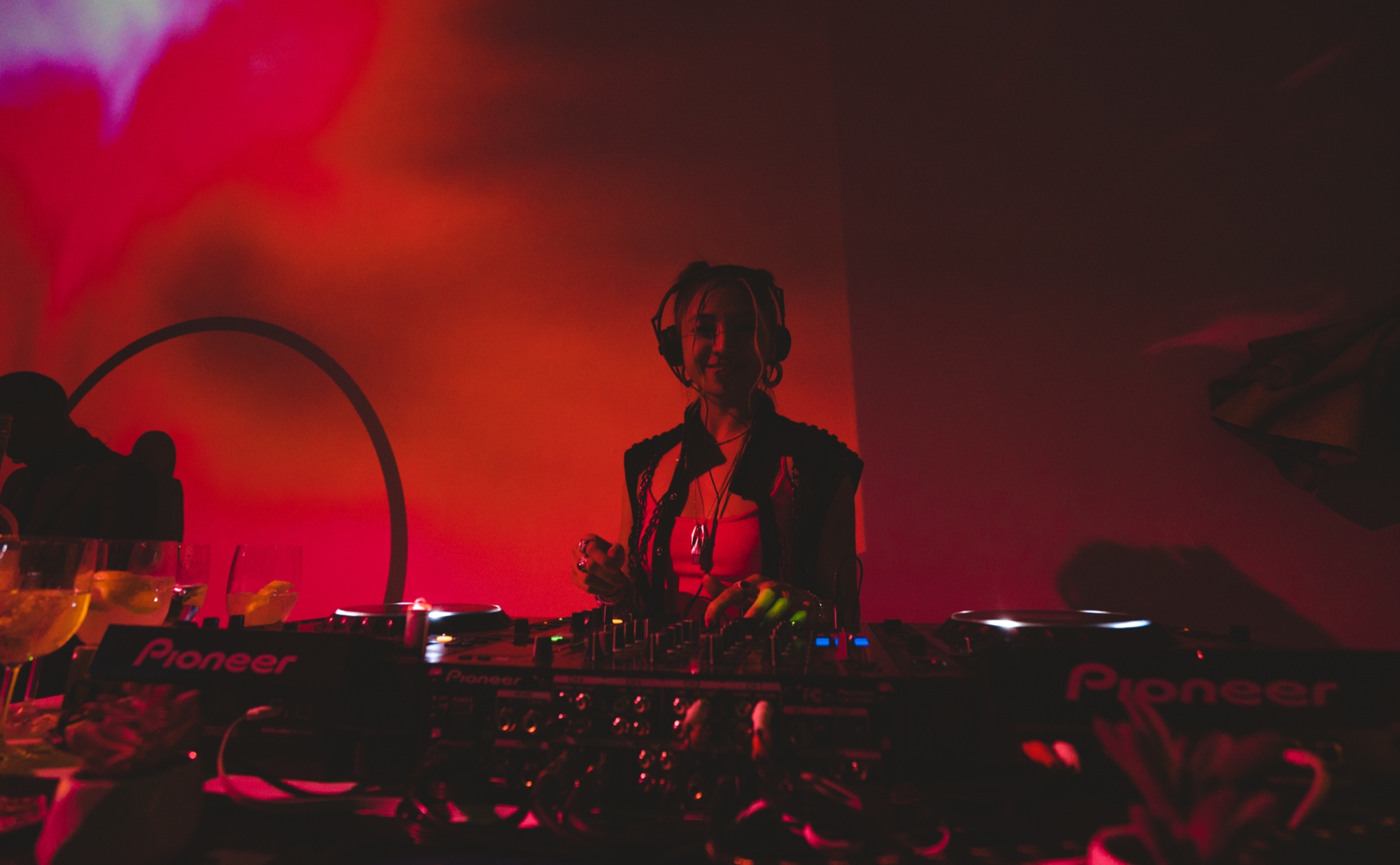 A DJ in headphones plays on Pioneer decks under red and pink projected light with cocktail glasses in front