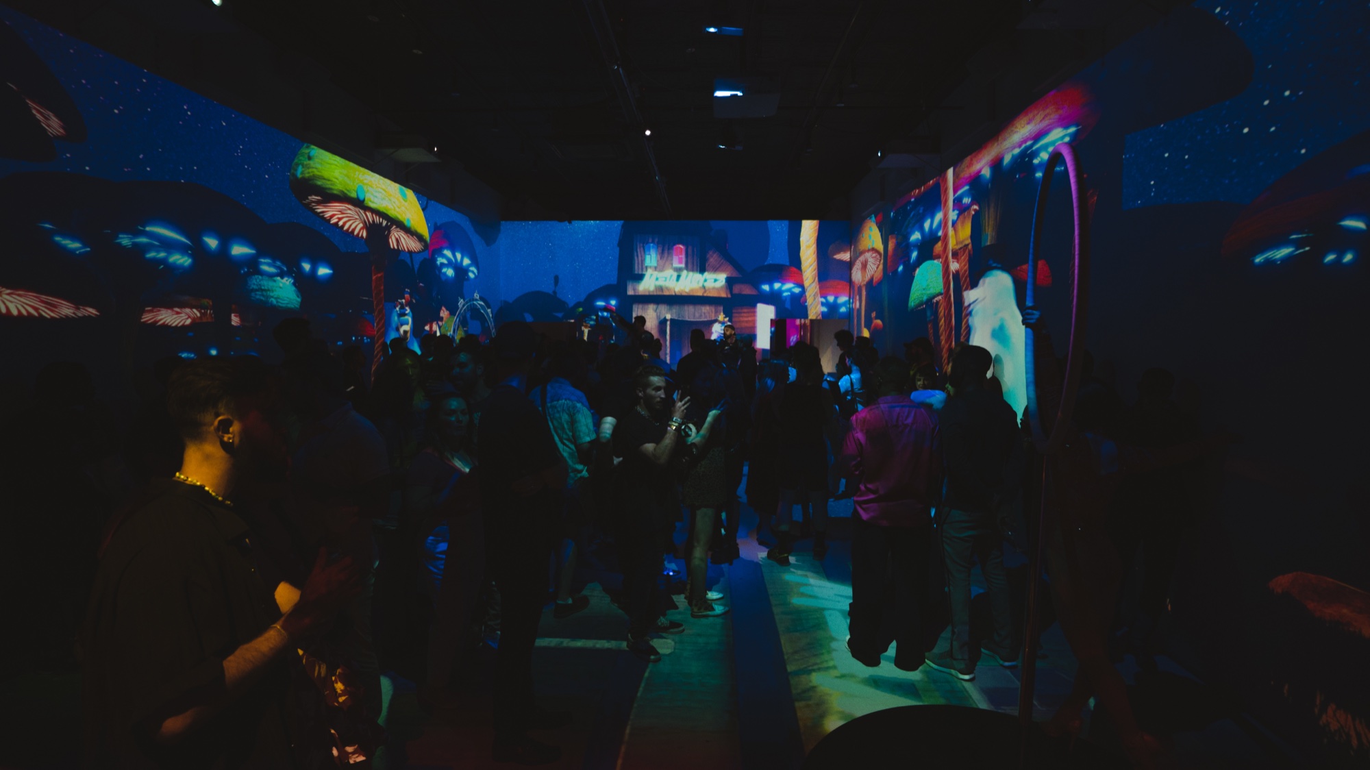 Silhouetted guests move through a dark hall under blue and purple projections of glowing mushrooms