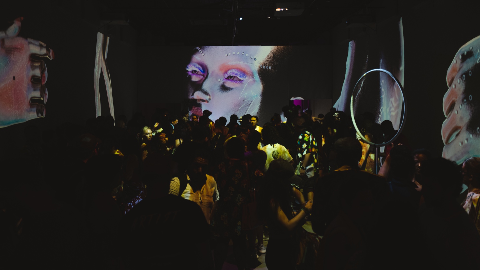 A crowd stands beneath a projected close up of a face with pink and blue makeup on a dark wall