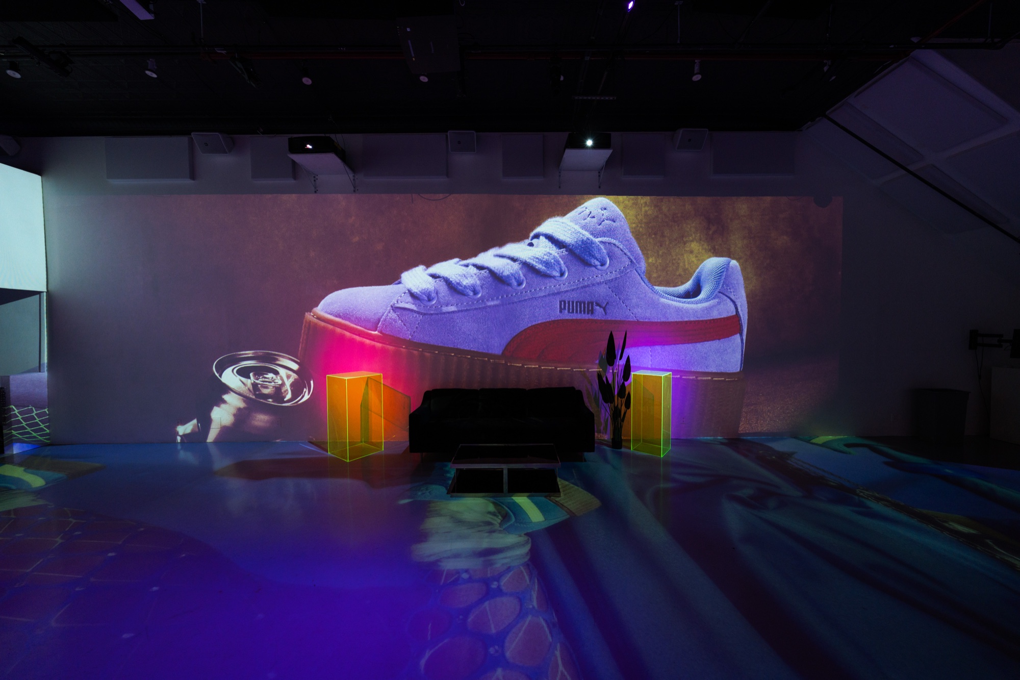 Giant projection of a white Puma platform sneaker on a wall above a black sofa and neon plinths
