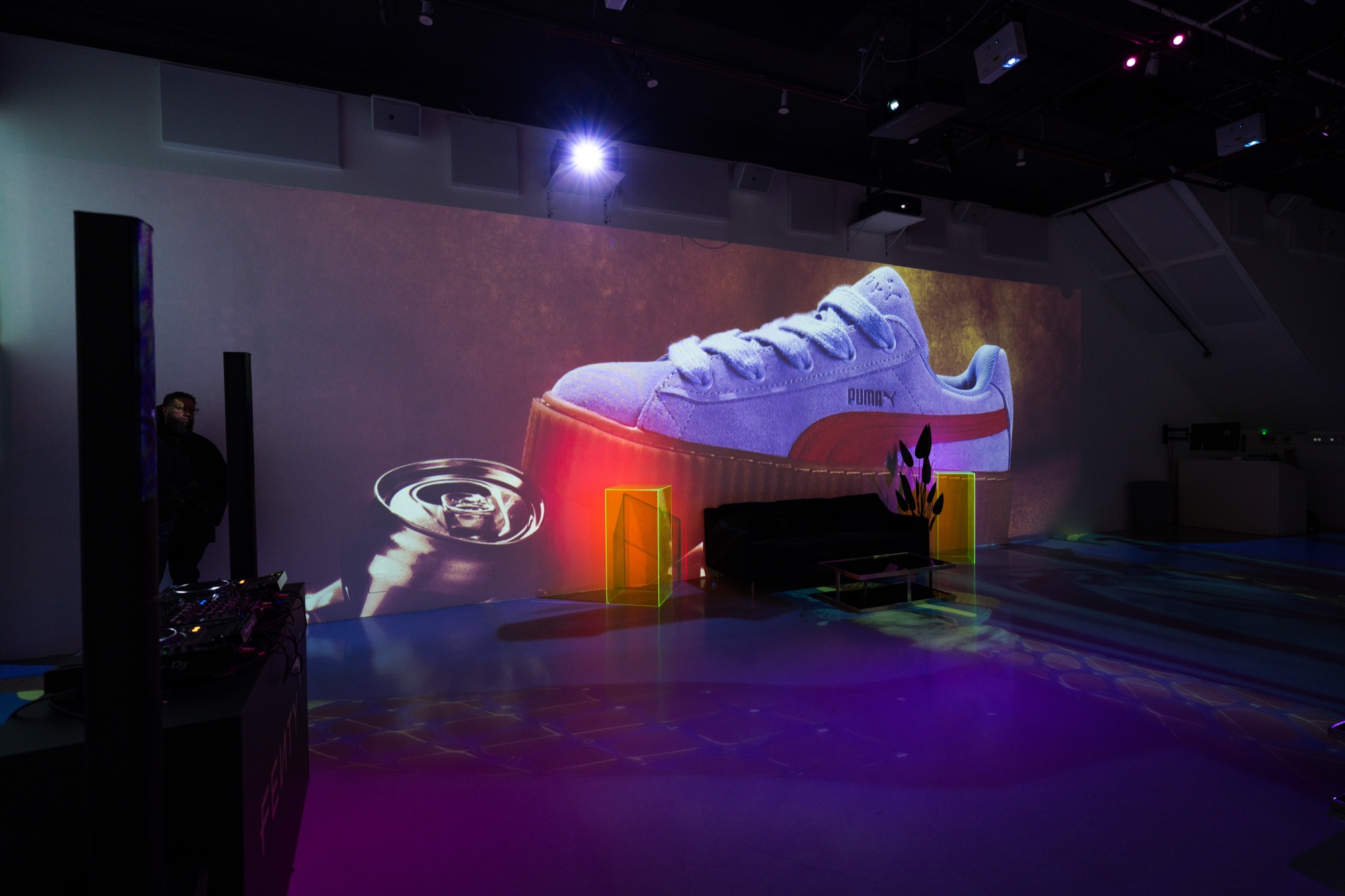 A giant white Puma sneaker projected on a wall above glowing yellow acrylic boxes on a purple lit floor