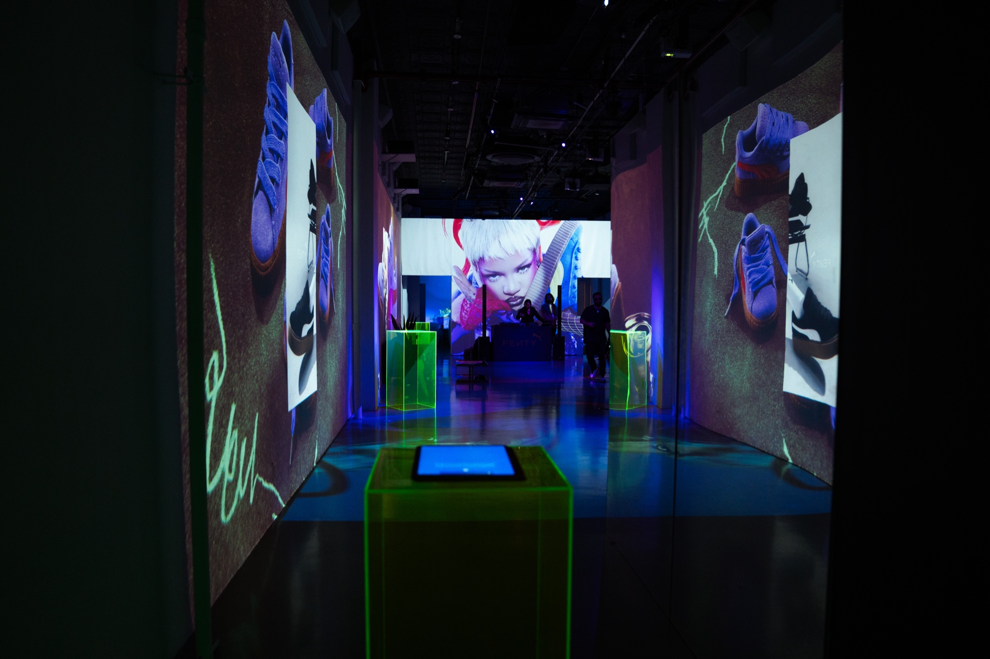 Dark corridor of projected sneaker imagery leading to a lit green acrylic plinth with a glowing screen