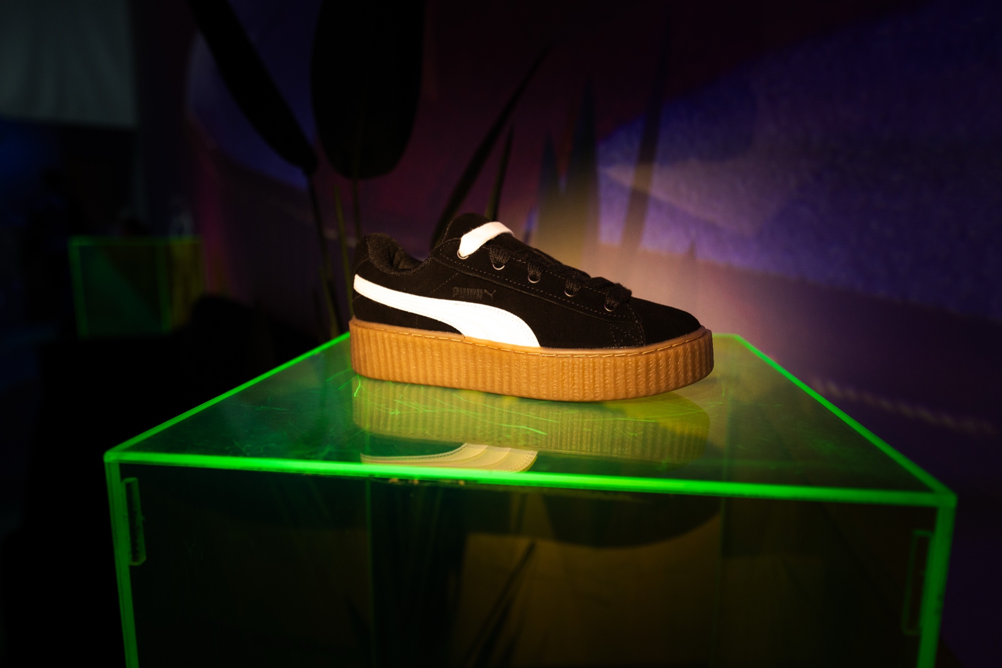 Black Puma suede creeper sneaker with a gum sole spotlit on a green edge lit acrylic plinth