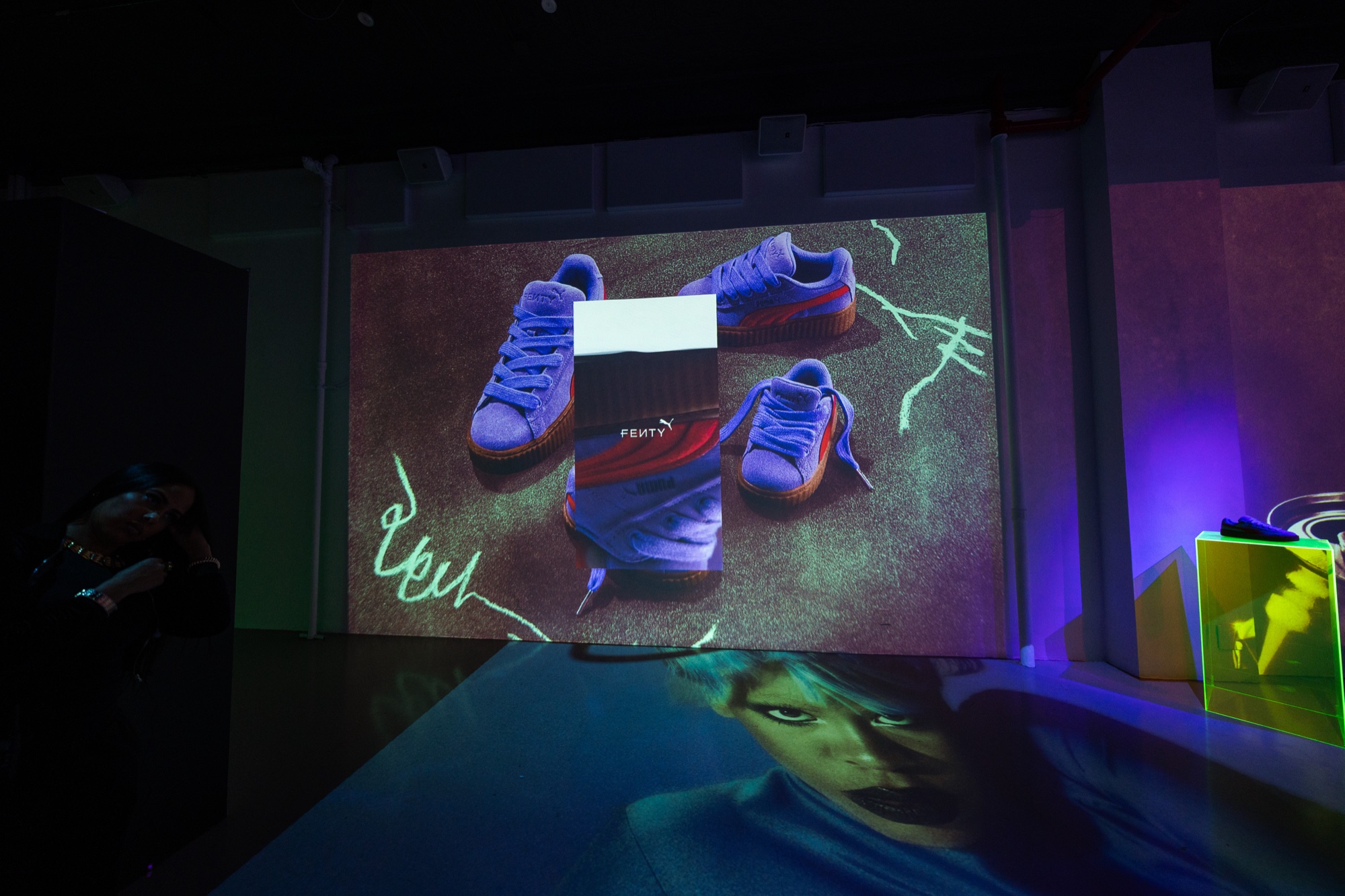Purple and orange sneakers beside a box are projected on a wall above a floor printed with a face