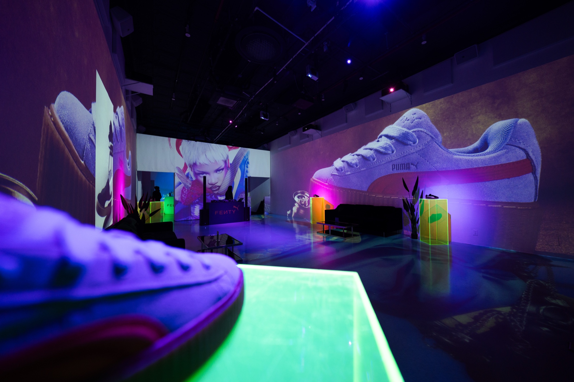 Low angle past a pale sneaker on a glowing green plinth toward projected Puma shoes in a purple lit room
